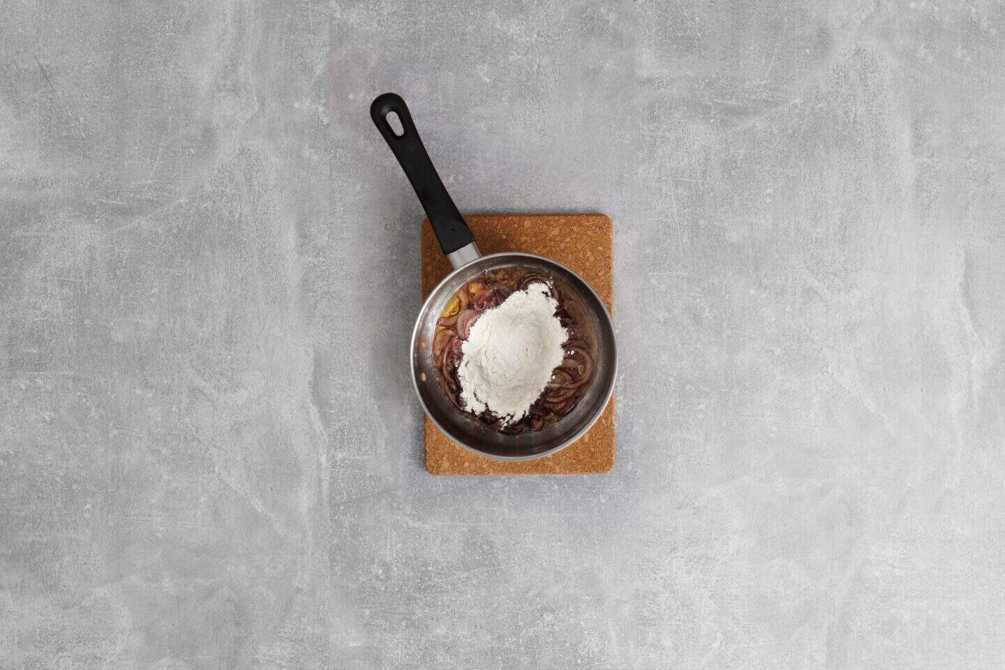 A saucepan with sliced onions and flour, perfect for starting Salisbury Steak with Rich Brown Gravy, sits on a cork mat on a gray countertop.