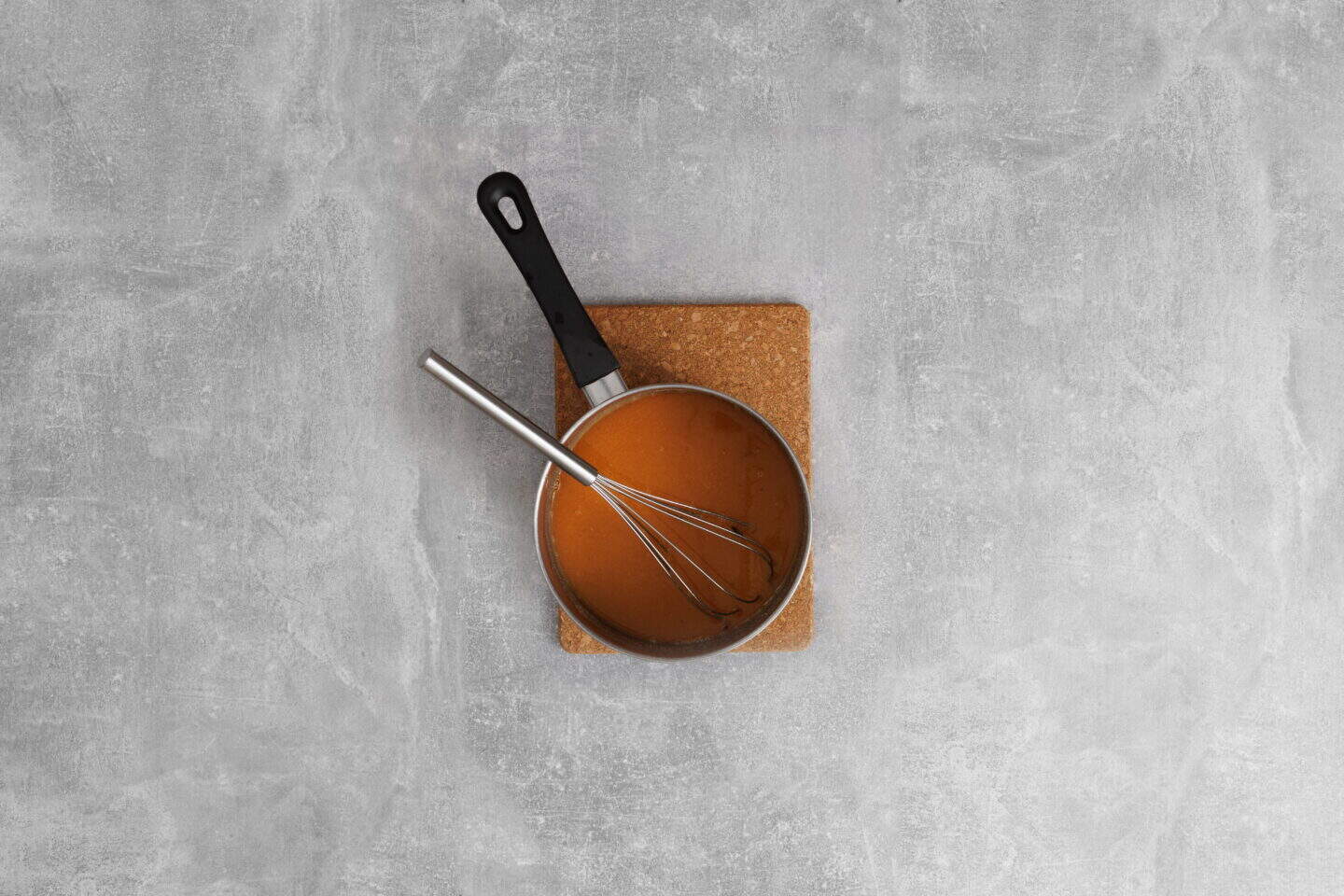 A saucepan filled with Salisbury Steak with Rich Brown Gravy and a whisk inside, placed on a cork mat on a gray textured surface.