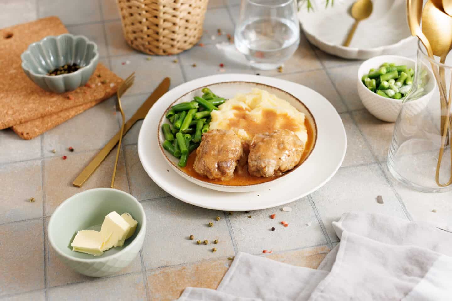 A plate of Salisbury Steak with Rich Brown Gravy, mashed potatoes, and green beans, surrounded by bowls of butter and extra green beans, cutlery, and a glass of water.