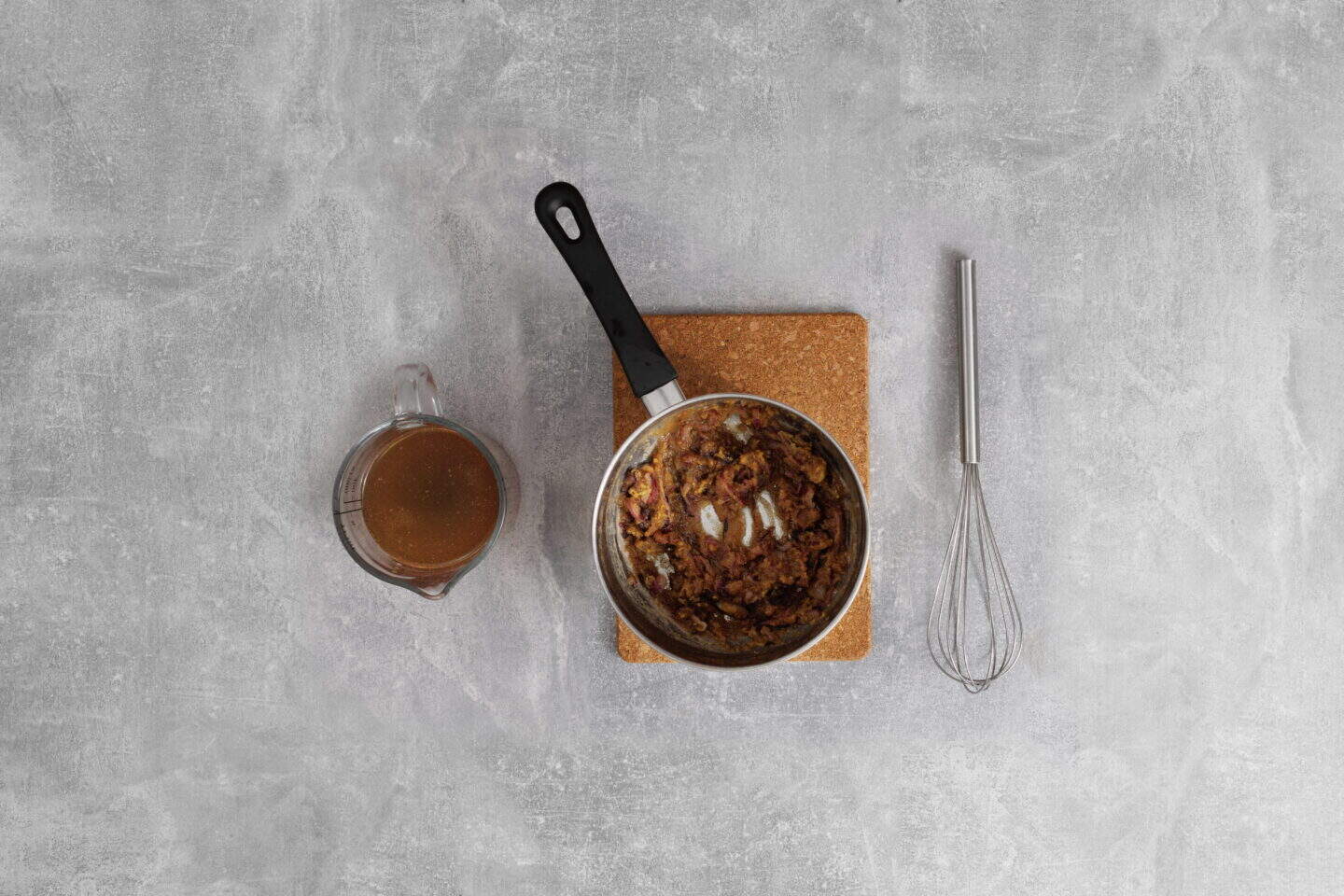 A pot with cooked onions sits on a cork mat next to a whisk and a measuring cup filled with Salisbury Steak with Rich Brown Gravy, all on a gray countertop.