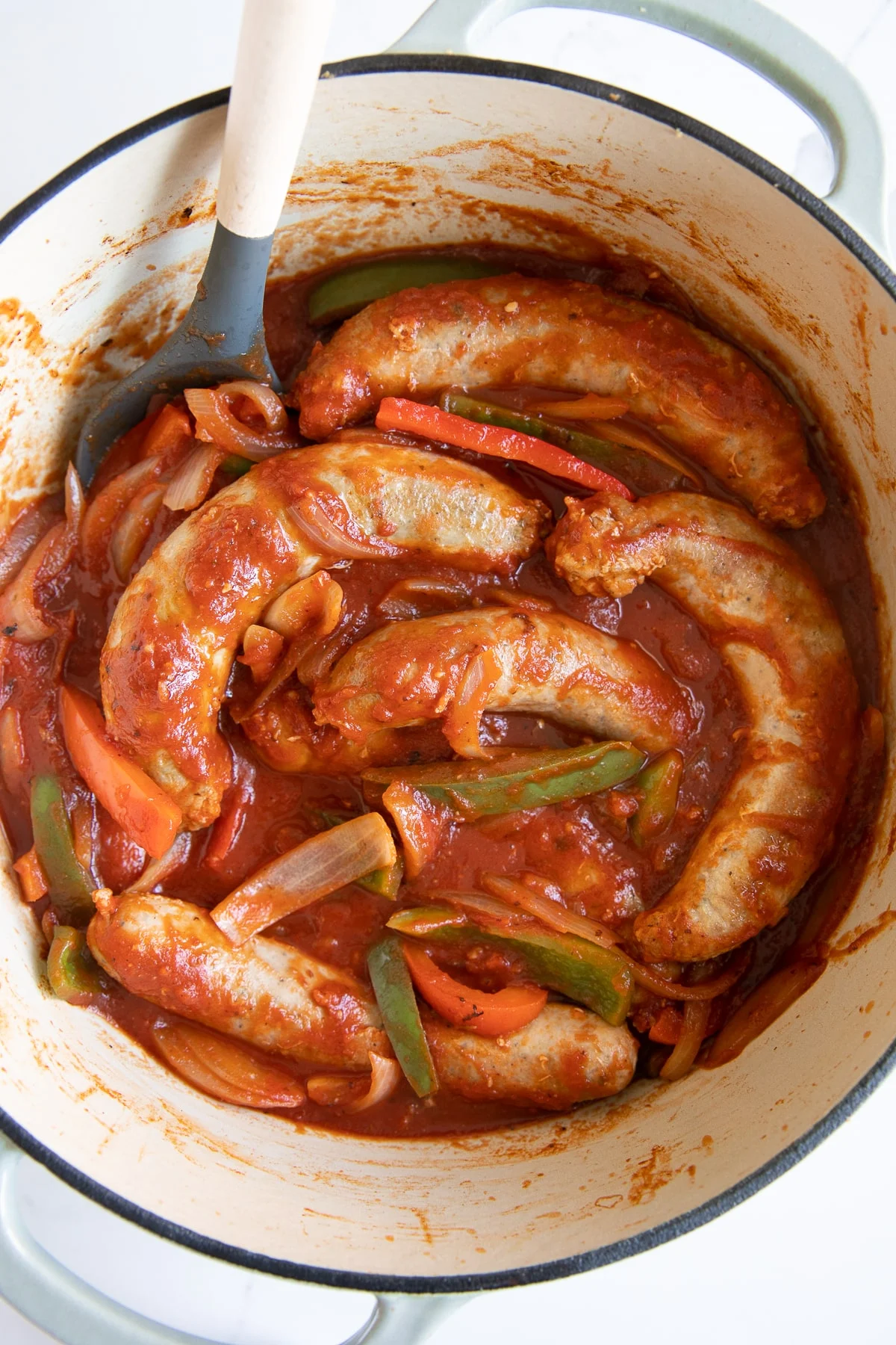 Sausages simmer in tomato sauce with sliced onions and bell peppers in a white Dutch oven, making this dish perfect for Italian potluck ideas, with a spoon resting on the side.