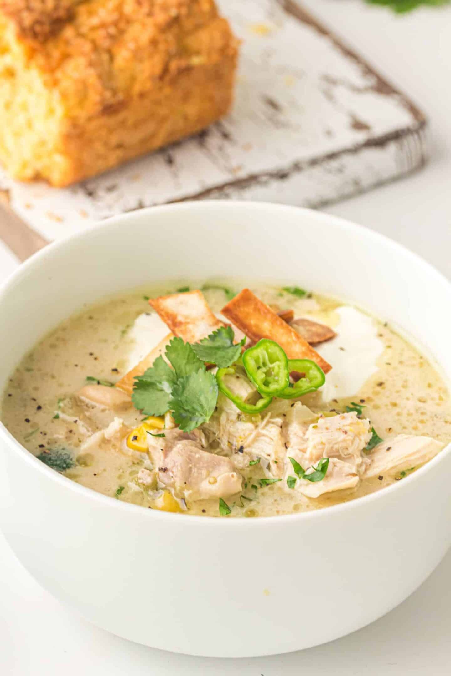 A bowl of creamy chicken soup garnished with tortilla strips, cilantro, and sliced green chili, with cornbread in the background—perfect for March dinner ideas.