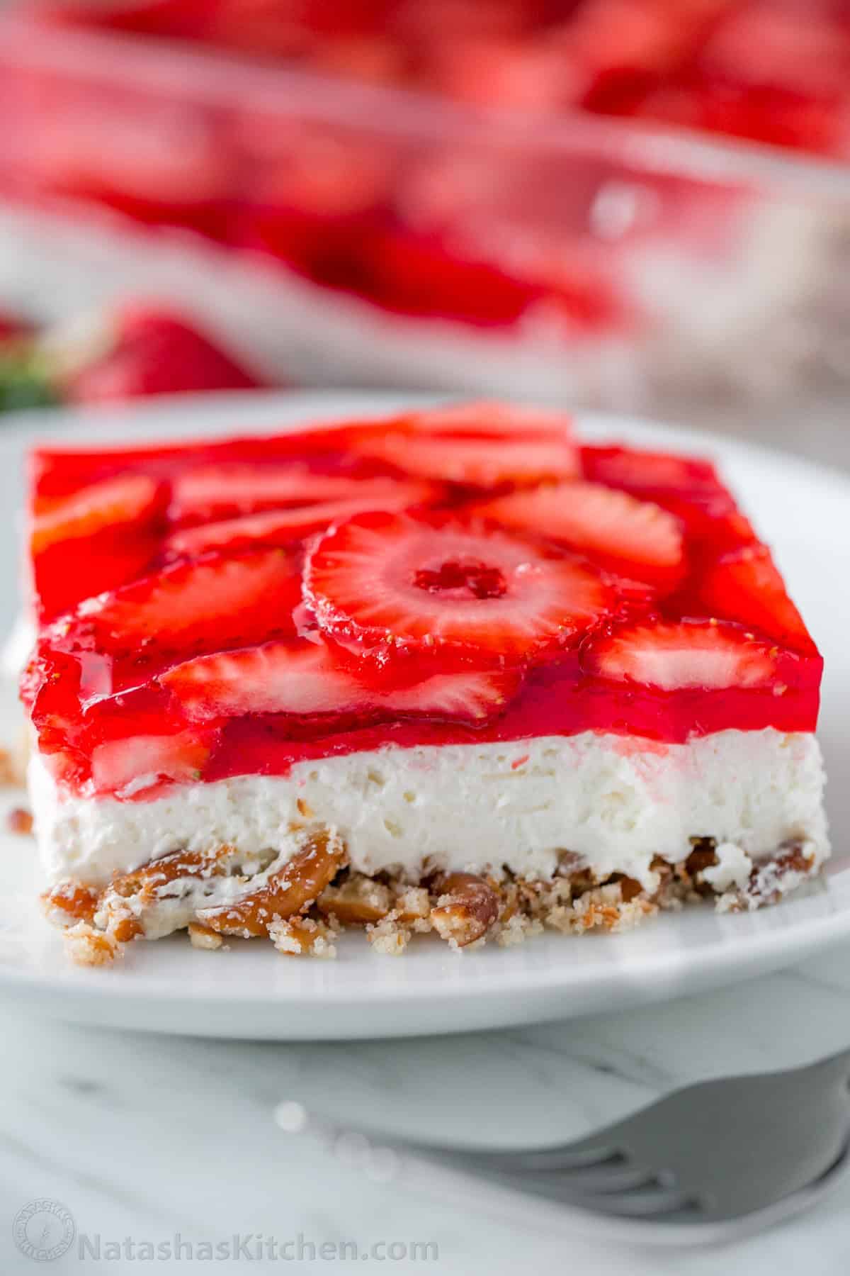A slice of layered dessert with a pretzel crust, creamy middle layer, and a top layer of strawberry slices in red gelatin—perfect for Easter recipes or other holiday side dishes—served on a white plate.