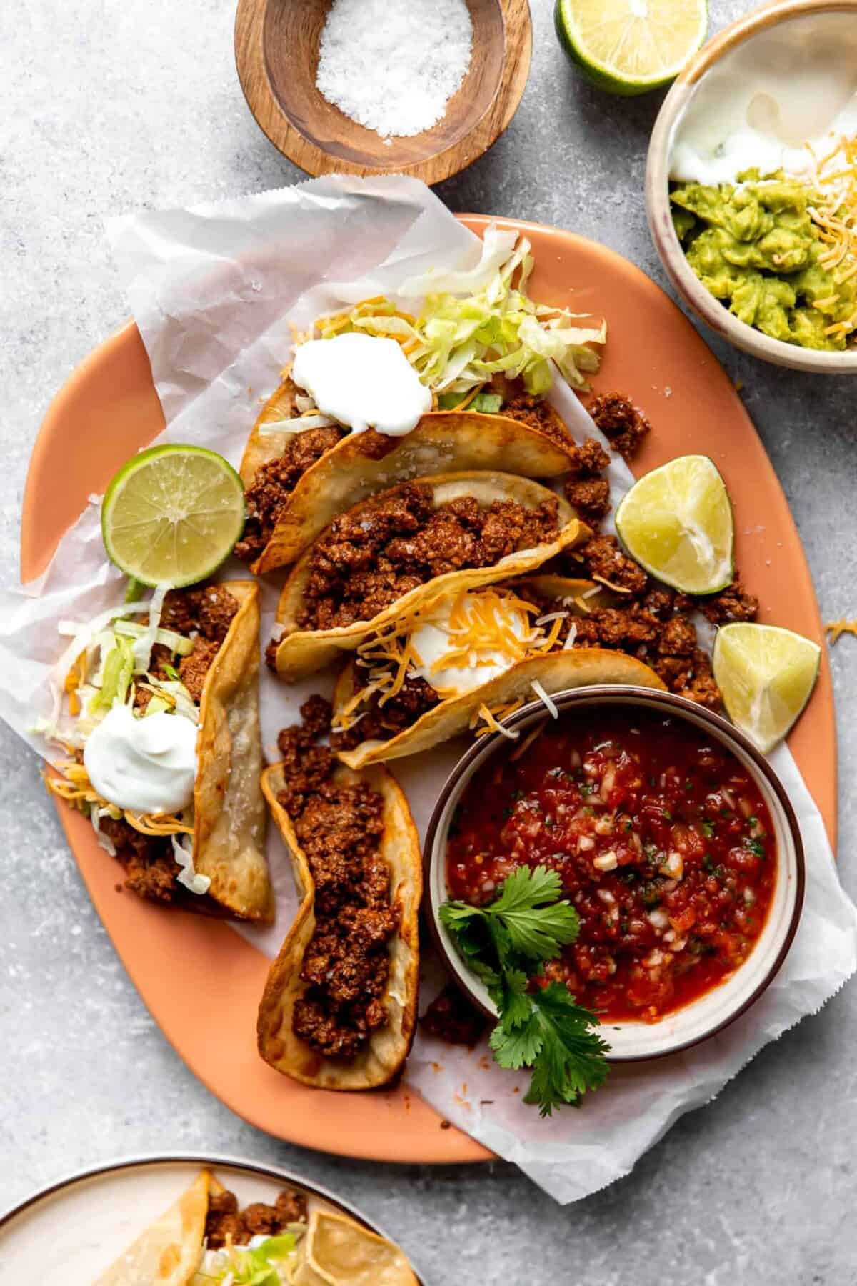 A plate of hard shell tacos filled with seasoned ground beef, topped with shredded lettuce, cheese, sour cream, and lime wedges—an easy choice for lazy dinner recipes—served with salsa, guacamole, and salt on the side.