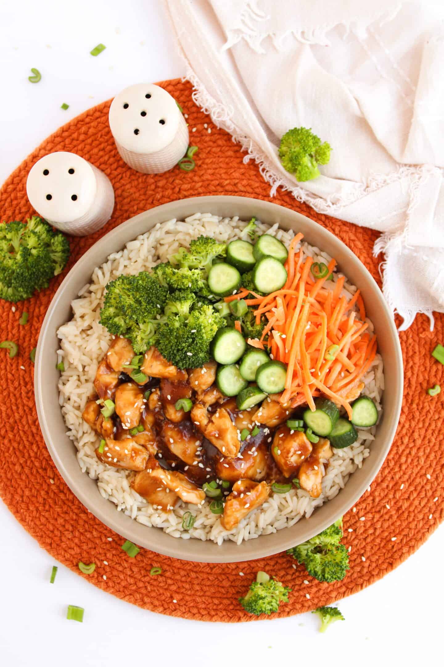 A bowl of rice topped with teriyaki chicken, broccoli, sliced cucumber, and shredded carrots makes the perfect Teriyaki Chicken Bowl, served on an orange mat with a napkin and salt and pepper shakers nearby.