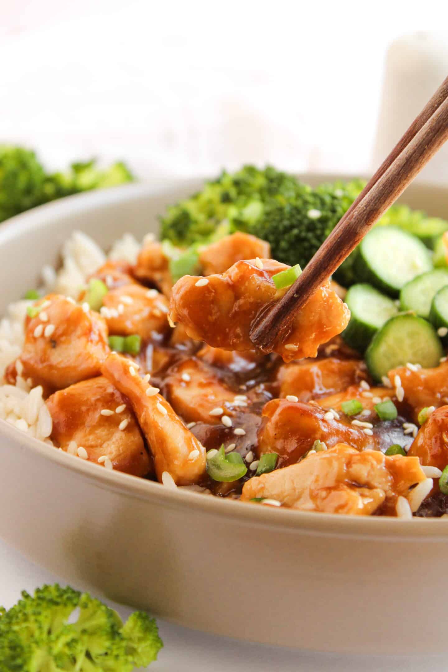 A bowl of rice topped with glazed Teriyaki Chicken, sliced cucumbers, broccoli, green onions, and sesame seeds, with chopsticks holding a piece of chicken—perfect for Teriyaki Chicken Bowls lovers.