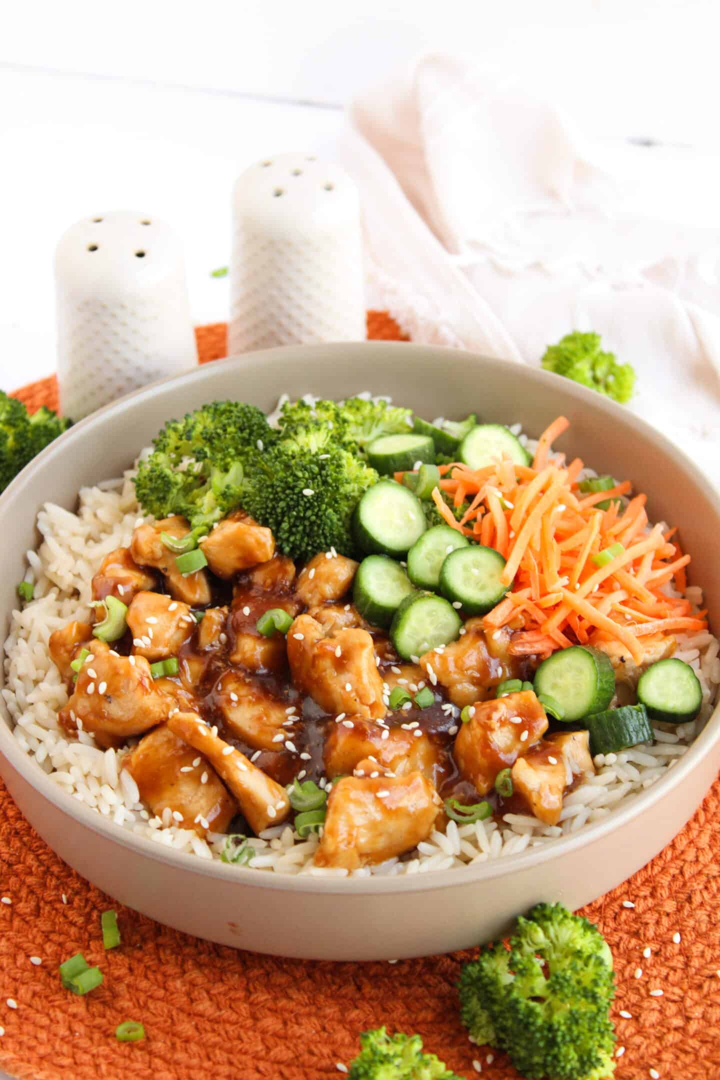A bowl of rice topped with teriyaki chicken, broccoli, sliced cucumbers, shredded carrots, green onions, and sesame seeds sits on an orange mat—perfect for fans of flavorful Teriyaki Chicken Bowls.