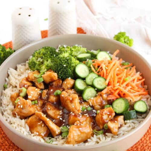 A bowl of rice topped with teriyaki chicken, broccoli, sliced cucumbers, shredded carrots, green onions, and sesame seeds sits on an orange mat—perfect for fans of flavorful Teriyaki Chicken Bowls.