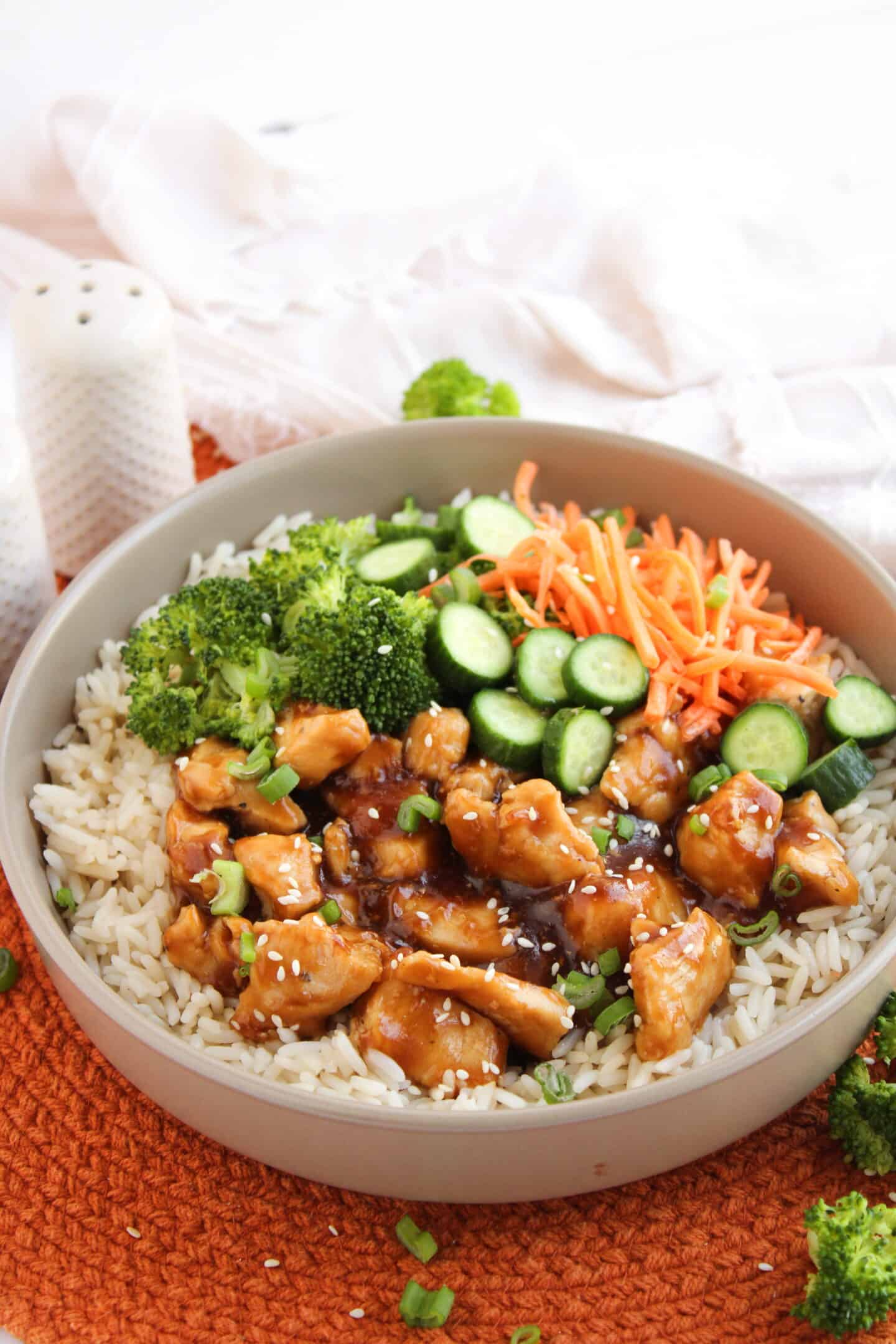 A bowl of white rice topped with glazed teriyaki chicken pieces, broccoli, sliced cucumber, shredded carrots, and sprinkled with sesame seeds and green onions—a delicious take on classic Teriyaki Chicken Bowls.