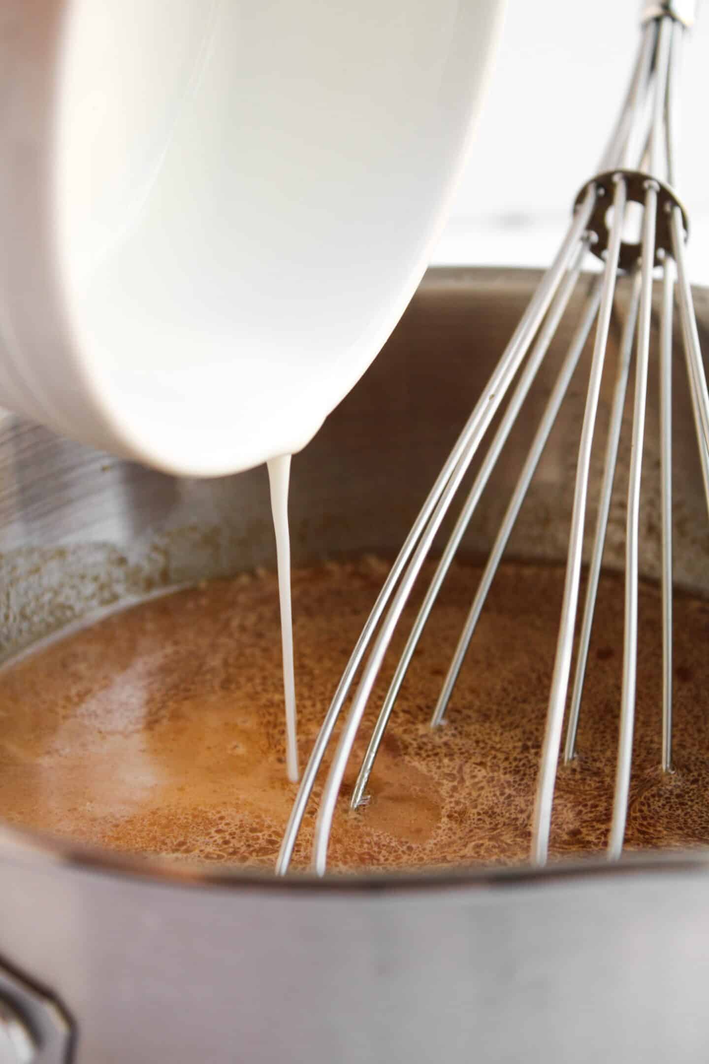 Milk is being poured from a white bowl into a saucepan with a brown liquid, possibly the sauce for Teriyaki Chicken Bowls, next to a whisk.