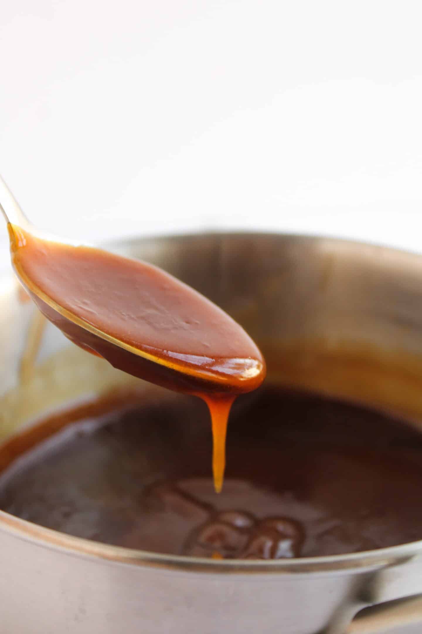 A metal spoon holds thick caramel sauce above a stainless steel pot filled with more sauce, perfect for drizzling over Teriyaki Chicken Bowls.