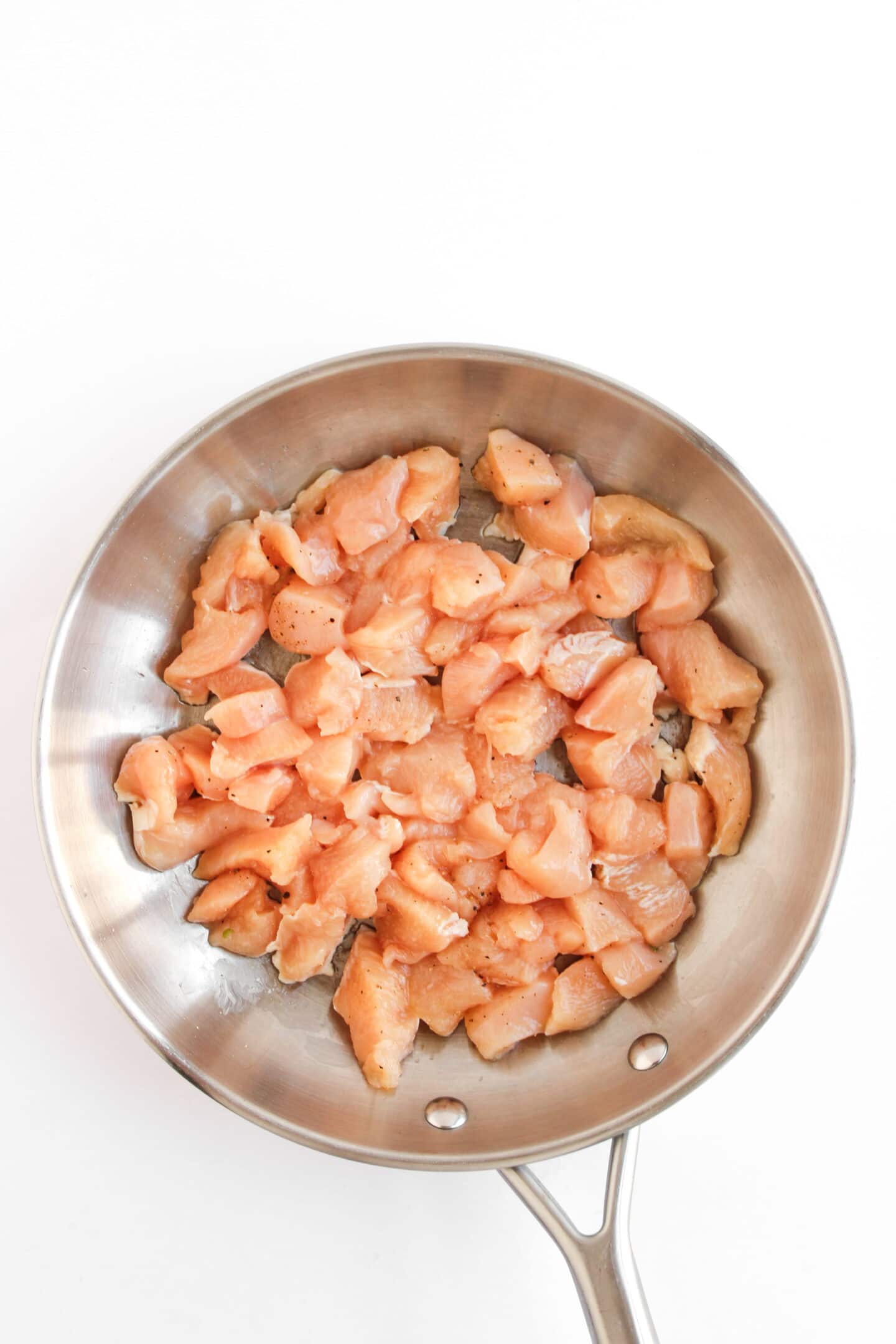 Diced raw chicken pieces in a stainless steel pan, ready to be cooked for Teriyaki Chicken Bowls, displayed on a white background.