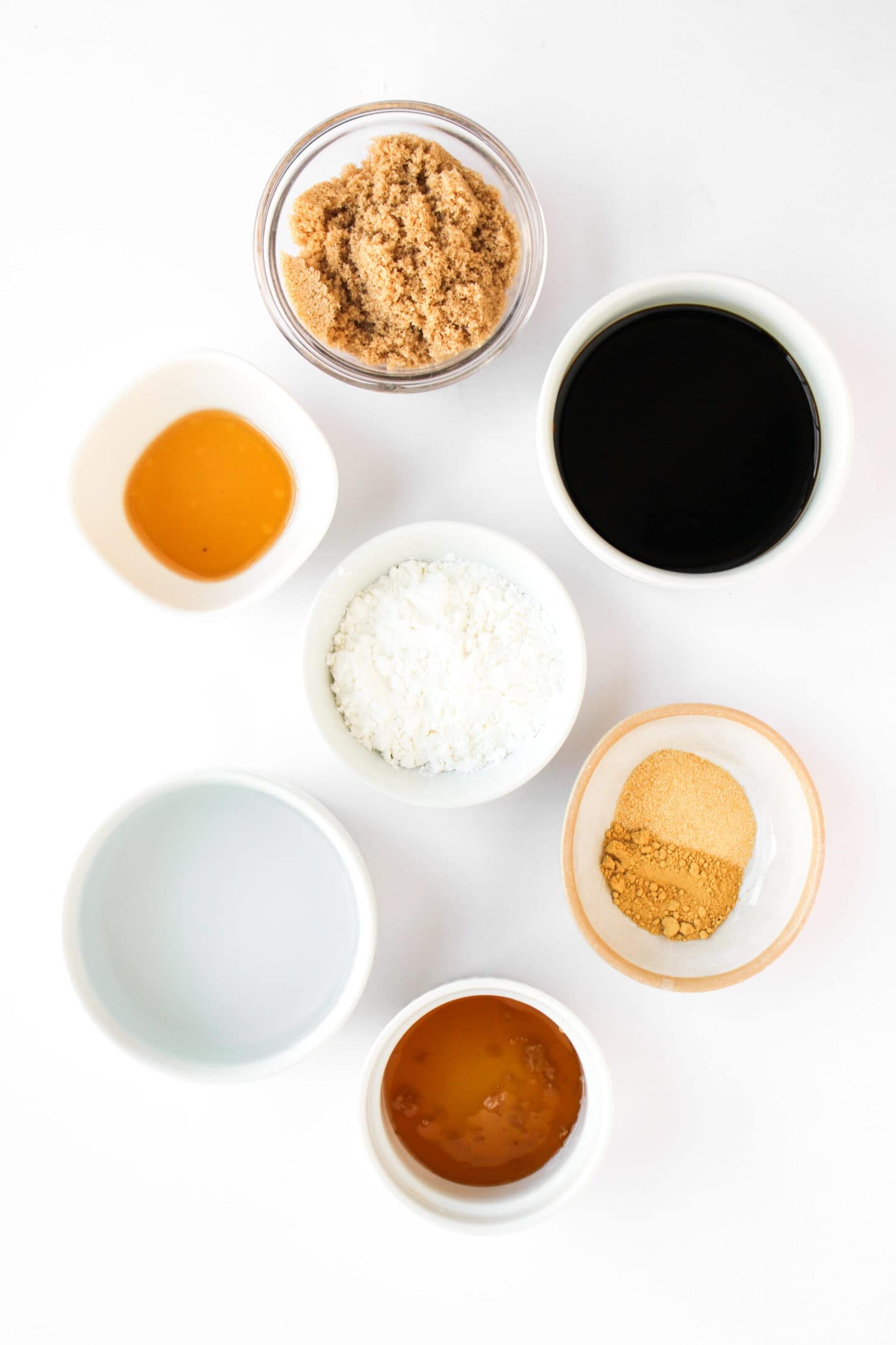 Seven small bowls containing ingredients like brown sugar, soy sauce, honey, and spices—perfect for making Teriyaki Chicken Bowls—are arranged on a white surface.