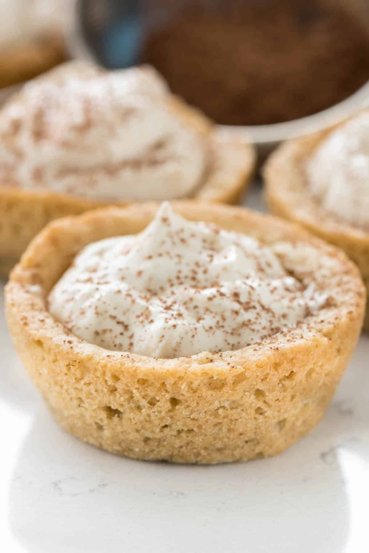 A cookie cup filled with whipped cream and dusted with cocoa powder sits on a white surface—an irresistible addition to your list of Italian potluck ideas, with similar desserts blurred in the background.
