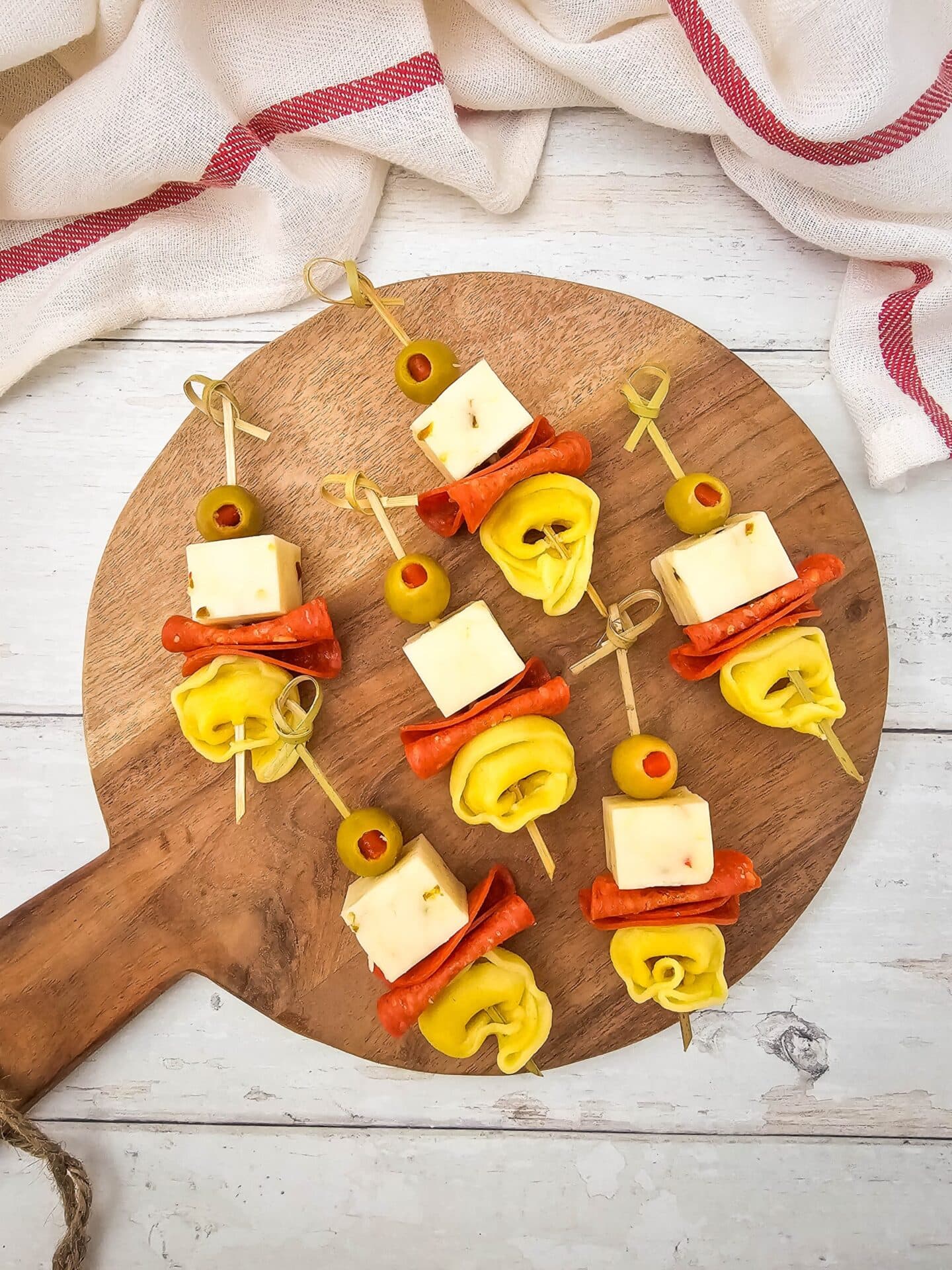 Skewers with cubes of cheese, slices of pepperoni, folded tortellini, and green olives arranged on a round wooden board—perfect Italian potluck fare and a crowd-pleaser among potluck dishes.