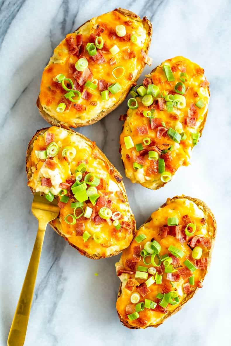 Four baked potato halves topped with melted cheese, bacon bits, and chopped green onions are arranged on a marble surface with a gold fork lifting one—perfect for serving as delicious Easter Side Dishes.
