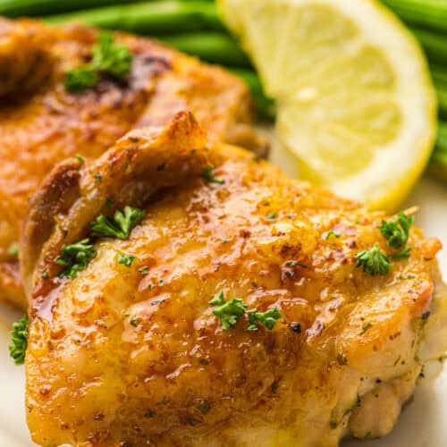 Close-up of air fryer chicken thighs garnished with parsley, served with a lemon wedge and asparagus in the background.