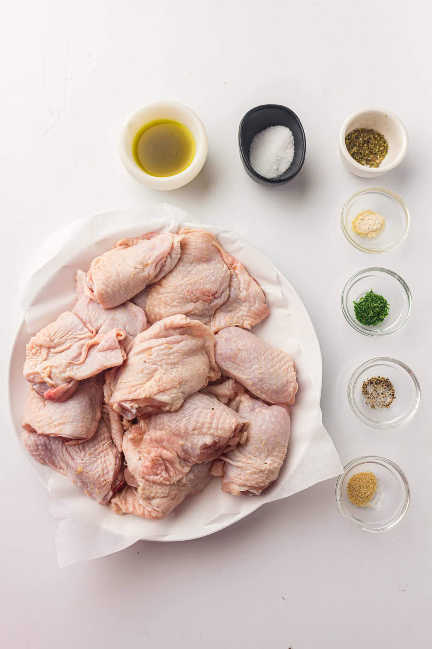 A plate of raw chicken thighs on paper is surrounded by small bowls of oil, salt, herbs, and spices on a white surface—perfect for preparing delicious air fryer chicken.