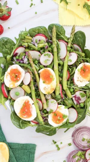 A fresh salad with spinach, radish, asparagus, peas, chopped red onion, and soft-boiled eggs, garnished with chives and black pepper on a white surface.