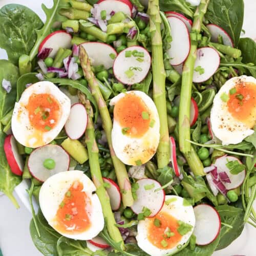 A fresh salad with spinach, radish, asparagus, peas, chopped red onion, and soft-boiled eggs, garnished with chives and black pepper on a white surface.