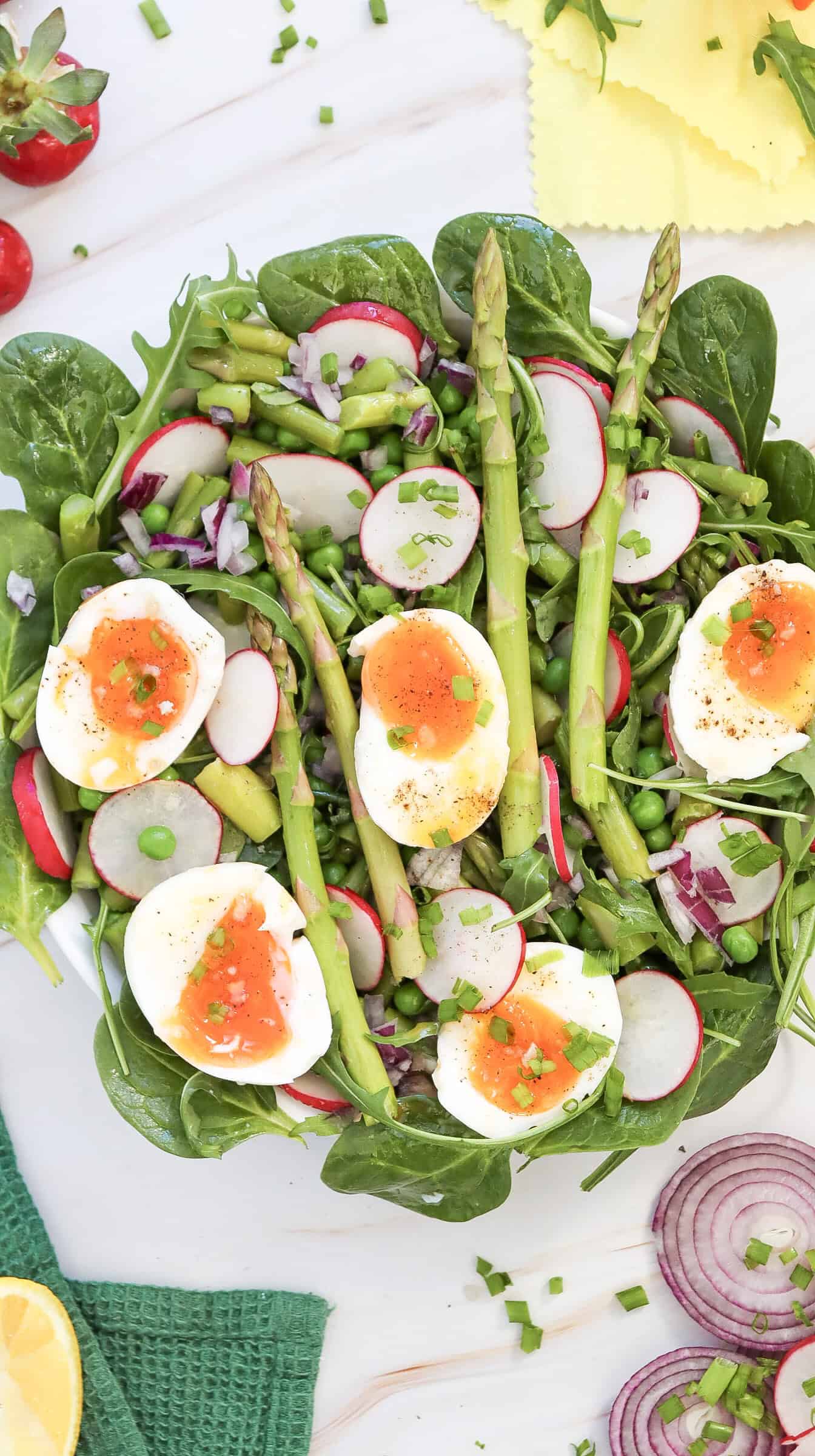 A fresh salad with spinach, radish, asparagus, peas, chopped red onion, and soft-boiled eggs, garnished with chives and black pepper on a white surface.
