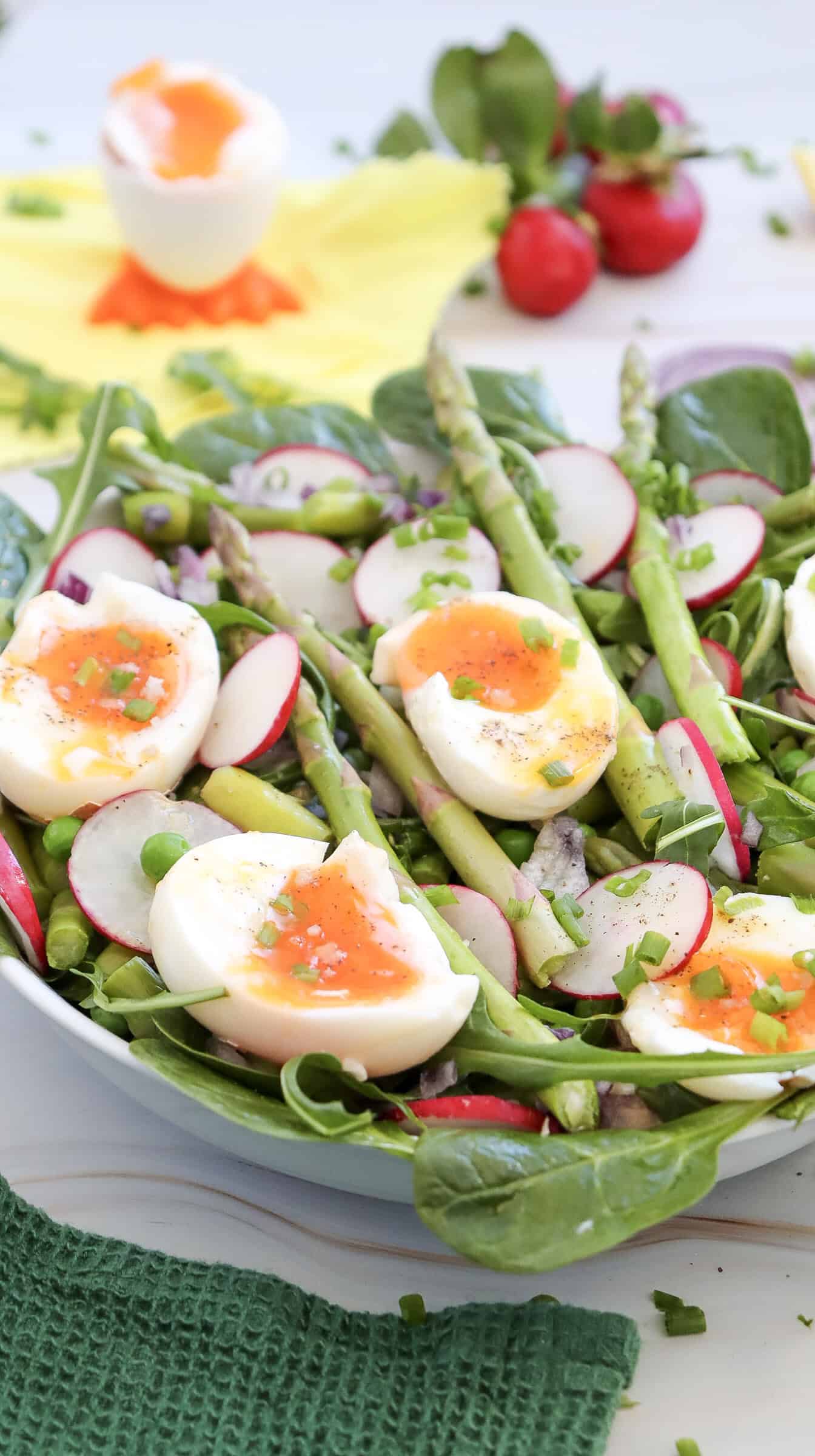 An asparagus salad with halved soft-boiled eggs, sliced radishes, spinach, and chopped chives, served in a white bowl and finished with a zesty lemon dressing. A soft-boiled egg and fresh radishes are in the background.