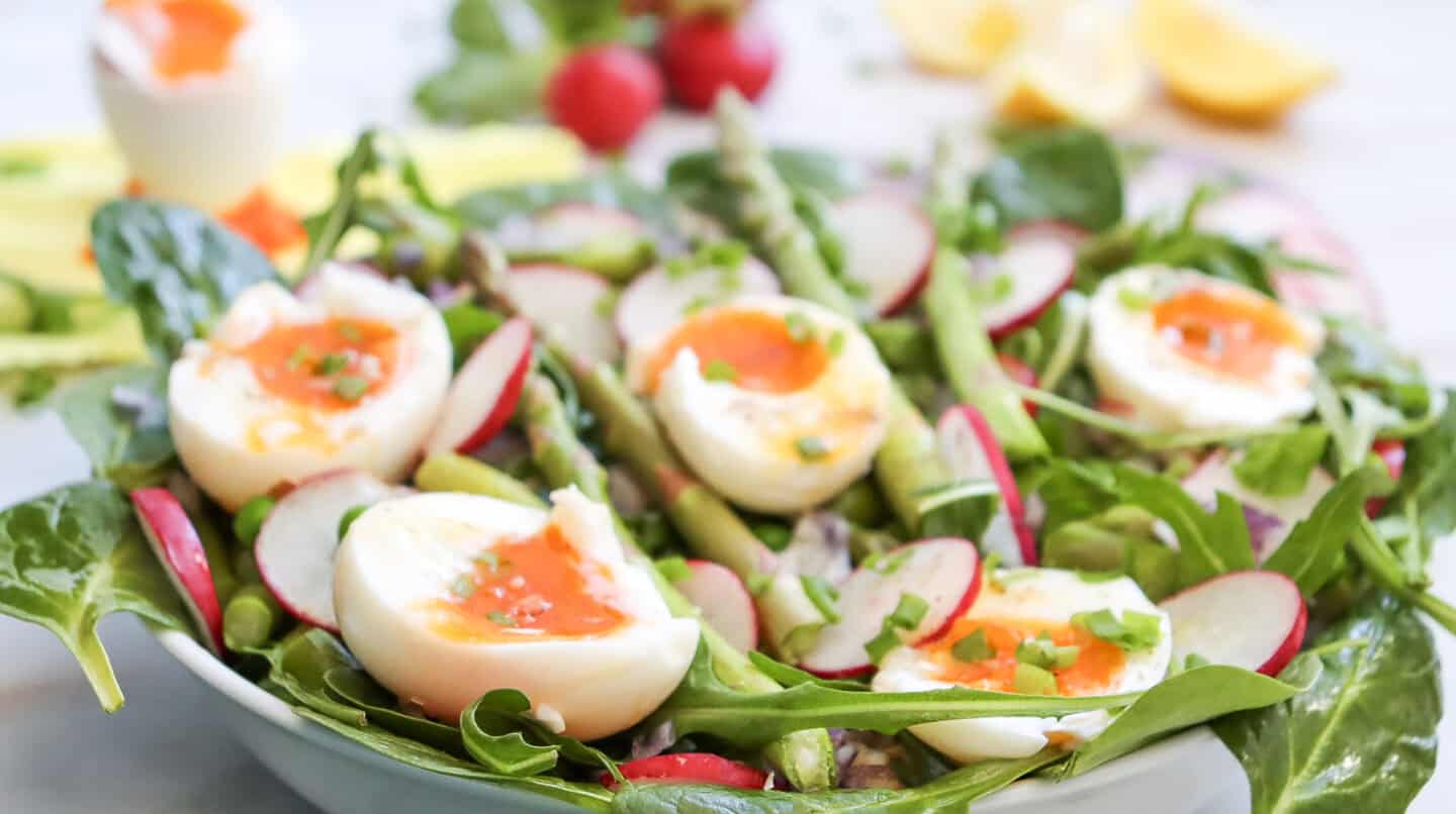 A salad with spinach, radishes, asparagus, and soft-boiled eggs, garnished with chopped herbs and drizzled with a zesty lemon dressing, served in a white bowl.