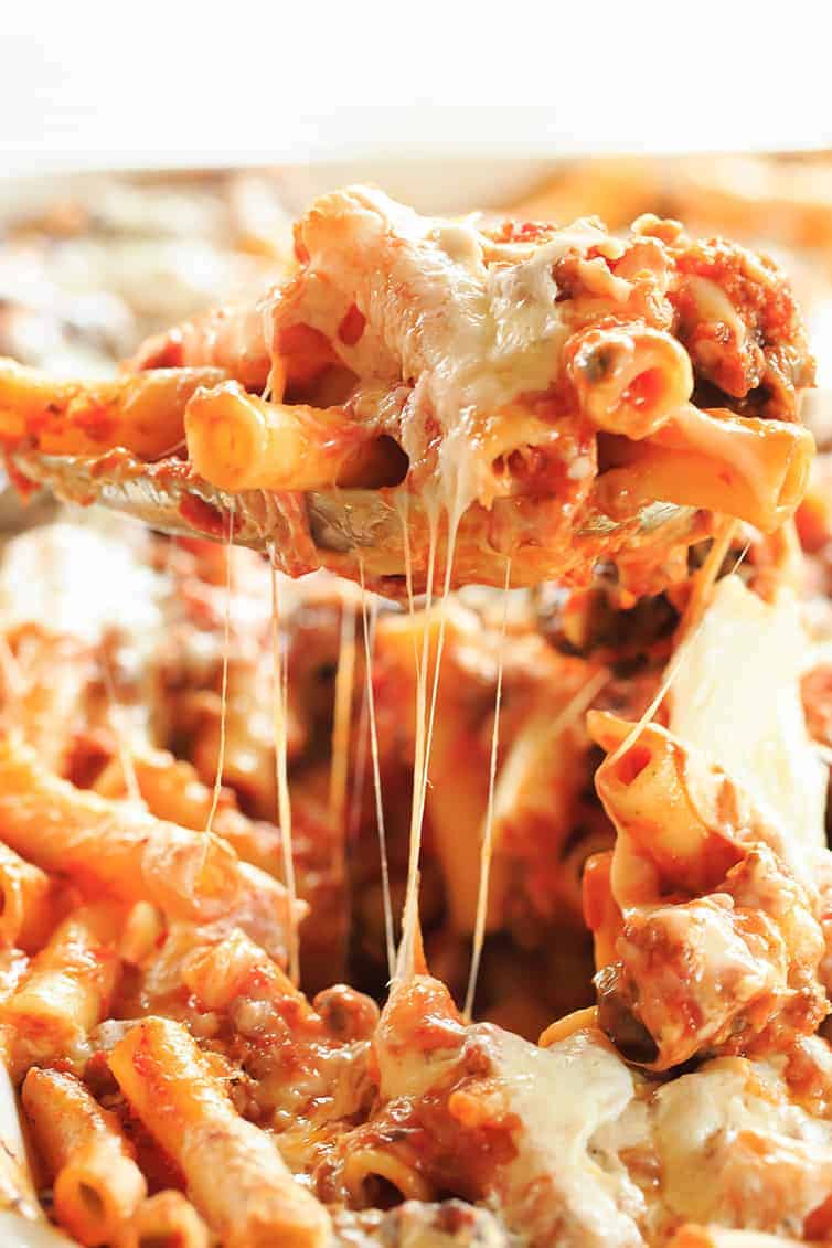 A serving of baked ziti is lifted from a cheesy, saucy casserole—perfect for an Italian potluck—with melted cheese stretching from the spoon to the pasta below.