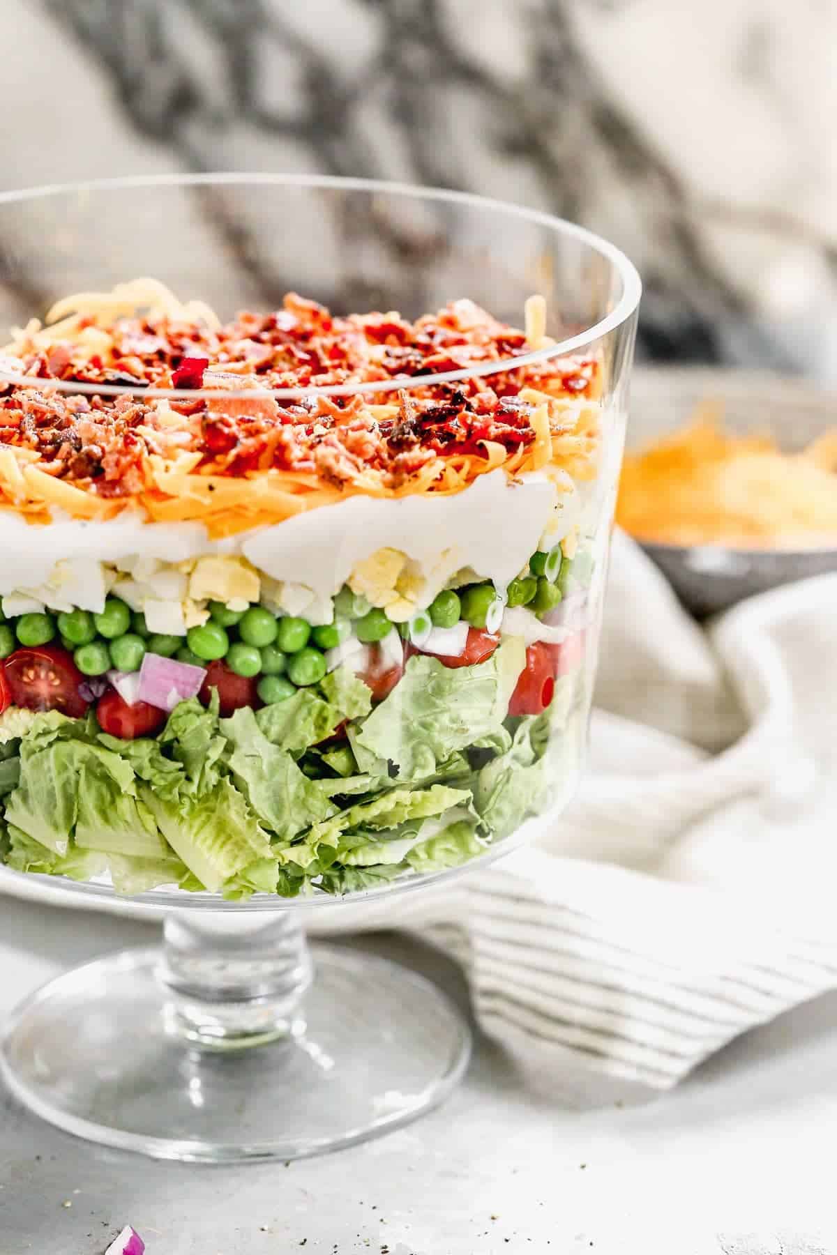 A glass trifle bowl displays a vibrant layered salad with lettuce, tomatoes, peas, eggs, cheese, bacon, and dressing—perfect as a colorful Easter Party Food centerpiece.