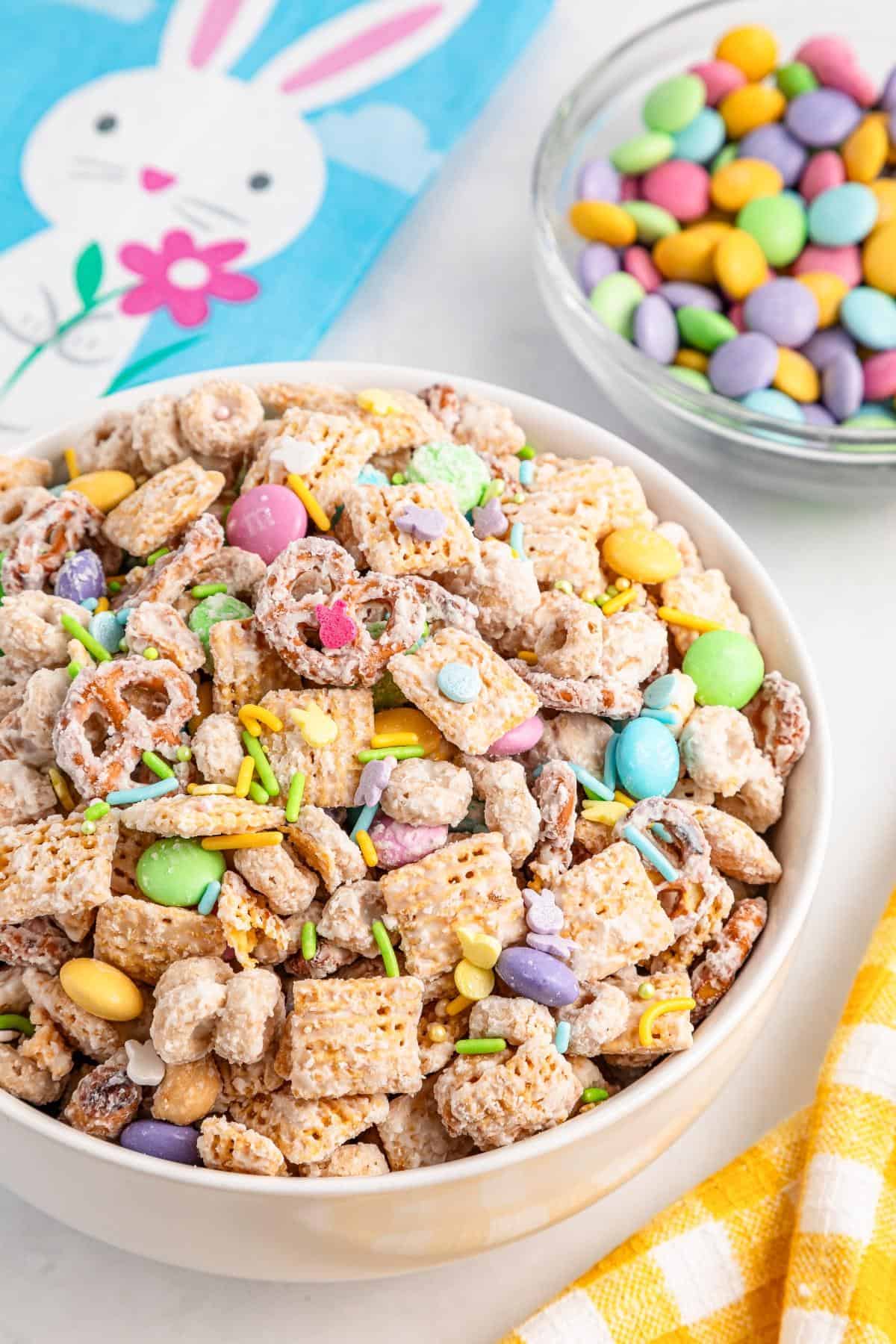 A bowl of sweet snack mix with cereal, pretzels, colorful candy, and sprinkles sits on a table next to a bowl of candy and a bunny napkin—perfect Easter Party Food for festive gatherings.
