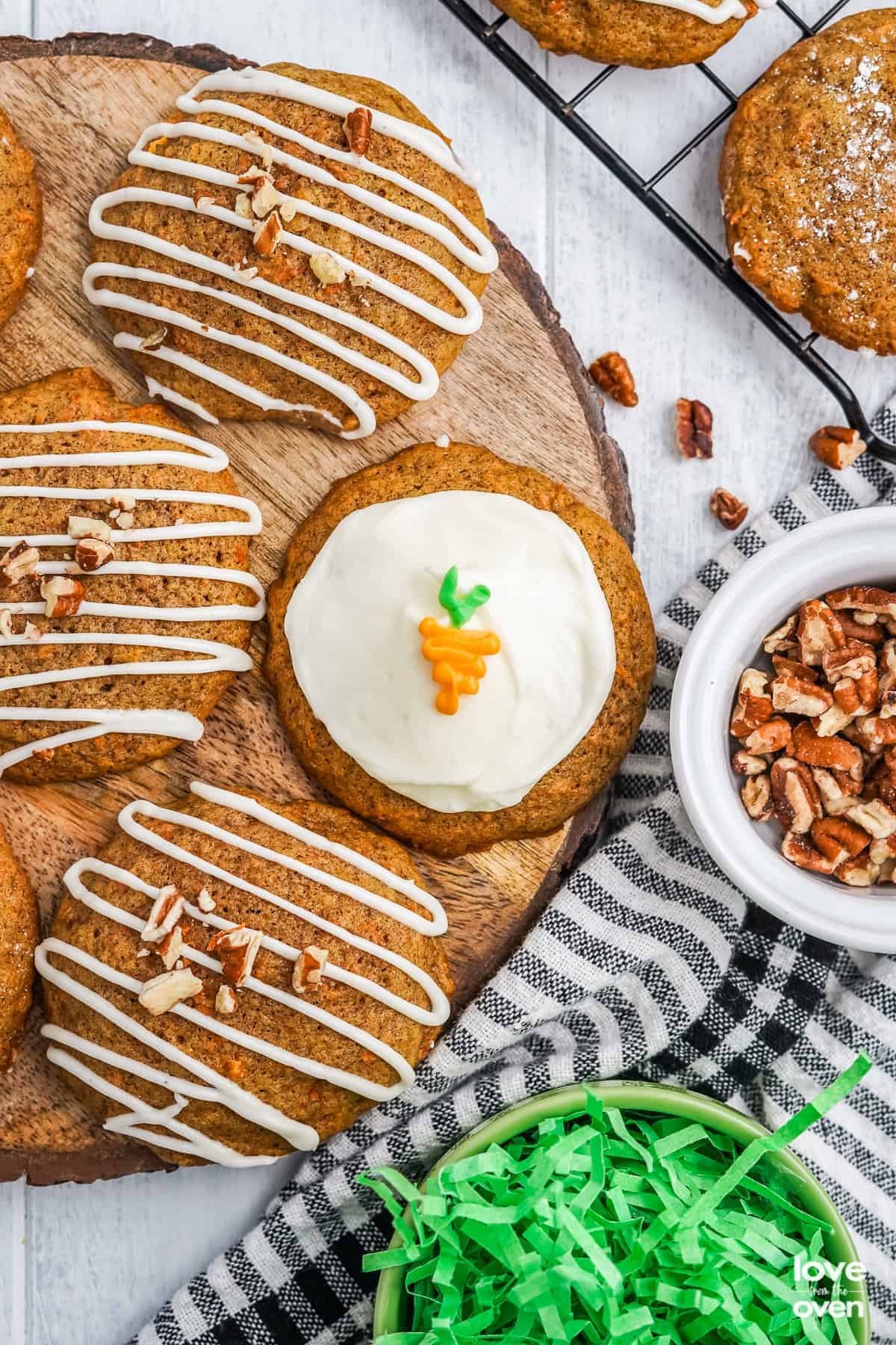 Several round cookies, some with white icing drizzle and chopped pecans, and one topped with white icing and a small carrot decoration, are arranged on a wooden board—perfect Easter Party Food alongside bowls of pecans and festive green paper grass.