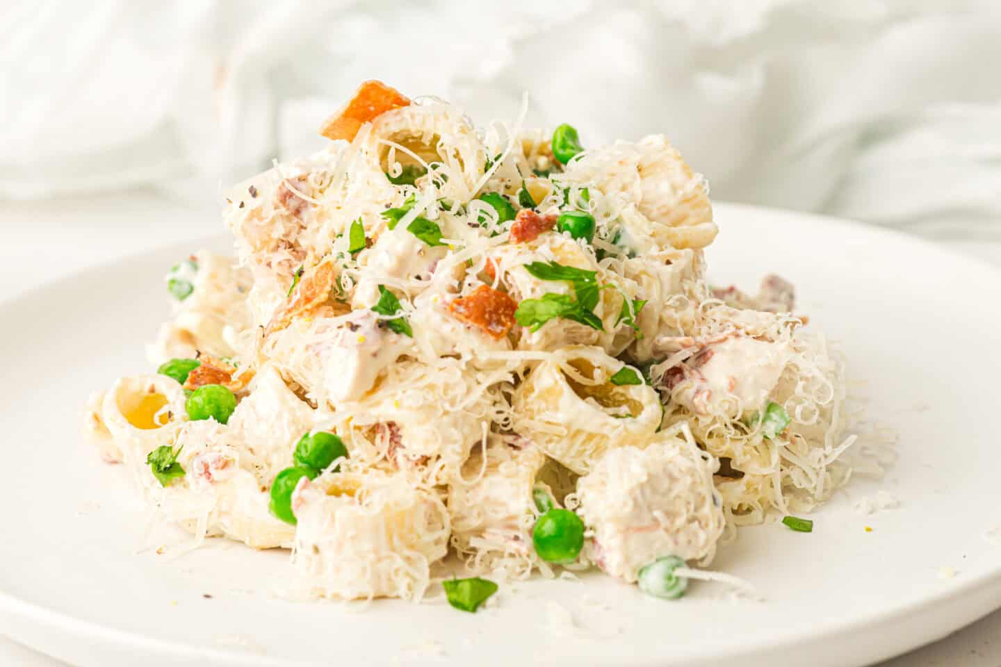 A plate of creamy chicken pasta salad with shredded cheese, green peas, chopped herbs, and savory pieces of bacon on a white background.