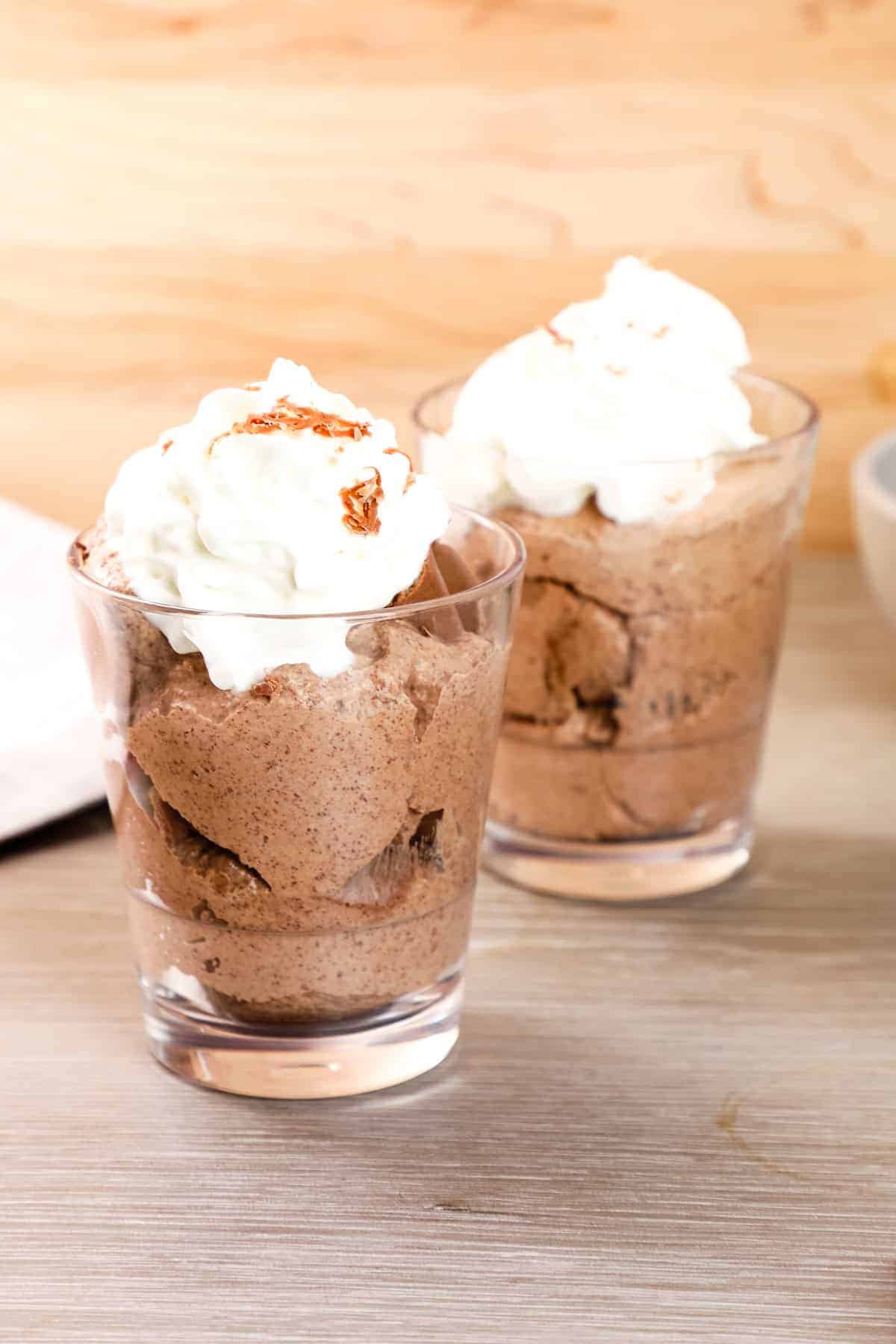 Two clear glasses filled with chocolate mousse, each topped with a dollop of whipped cream, make elegant Italian potluck ideas, beautifully presented on a wooden surface.