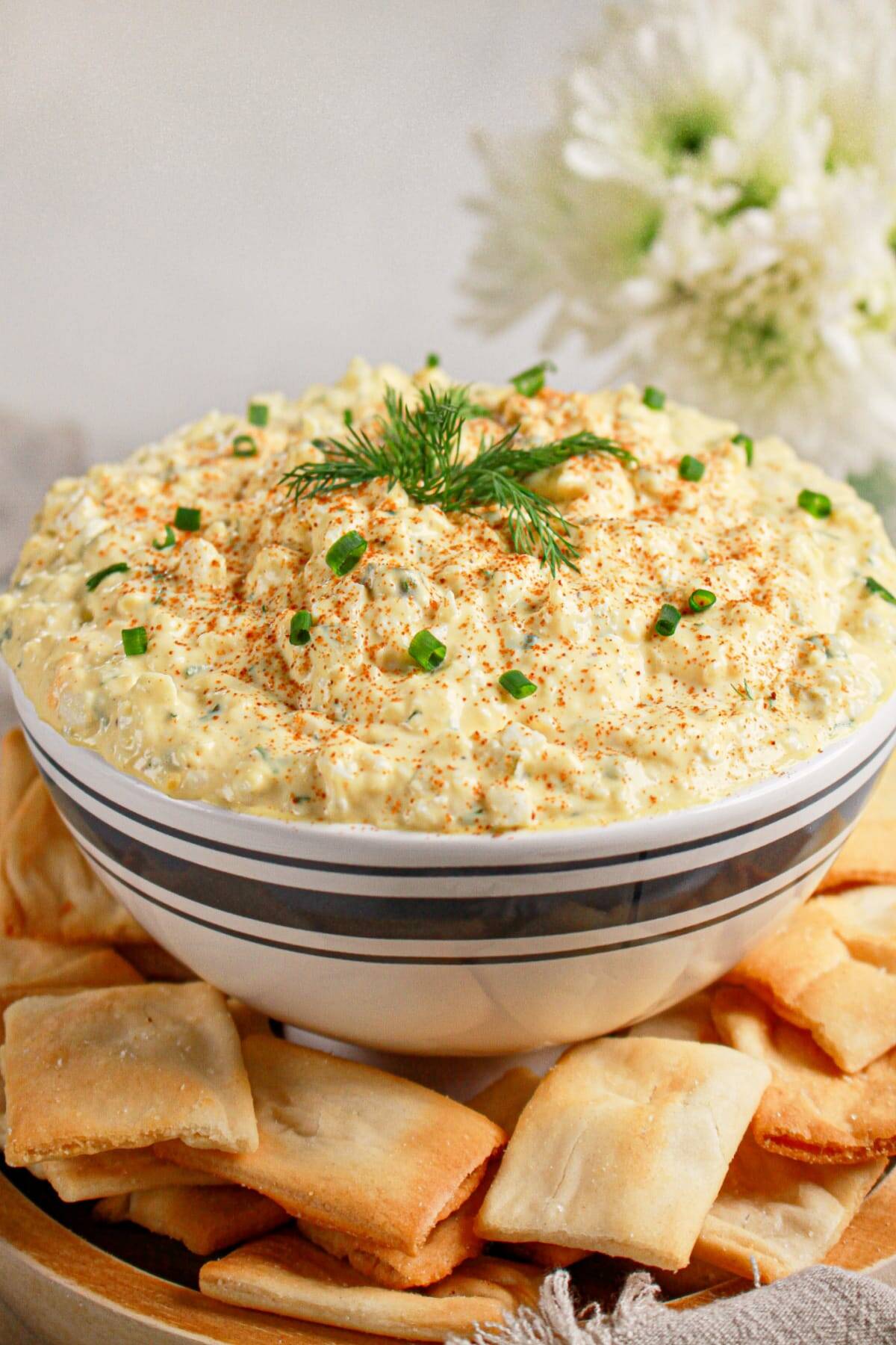 A bowl of creamy dip garnished with chopped chives and dill, surrounded by square crackers on a wooden tray—perfect for serving as Easter party food.