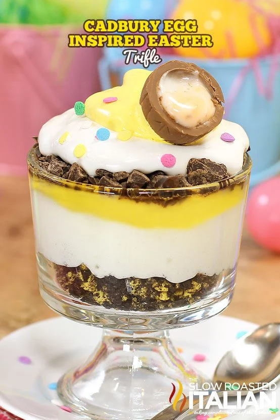 A layered trifle dessert in a glass, topped with whipped cream, yellow sauce, chocolate pieces, colorful sprinkles, and a halved chocolate egg—perfect as festive Easter party food.