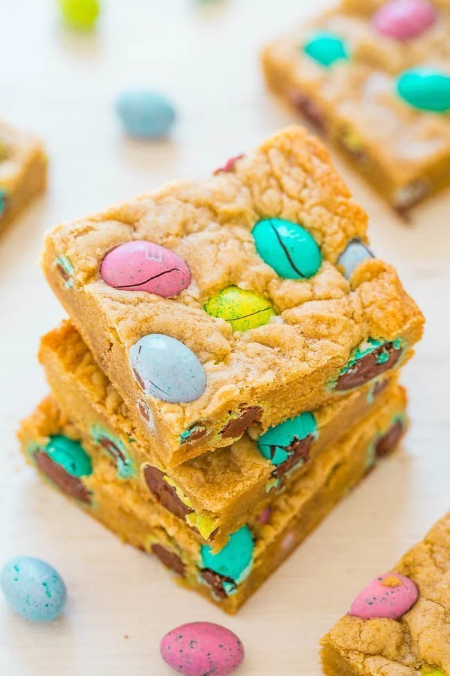 Three stacked cookie bars with colorful candy-coated chocolate eggs, perfect as Easter party food, with more bars and candies scattered on a light surface.