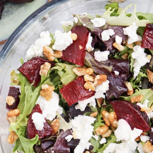 A glass bowl of salad featuring mixed greens, roasted beets, crumbled goatcheese, and walnuts, with plates of beets and goatcheese in the background.