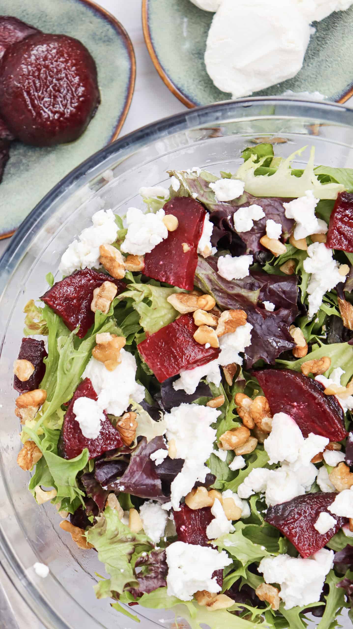A glass bowl of salad featuring mixed greens, roasted beets, crumbled goatcheese, and walnuts, with plates of beets and goatcheese in the background.