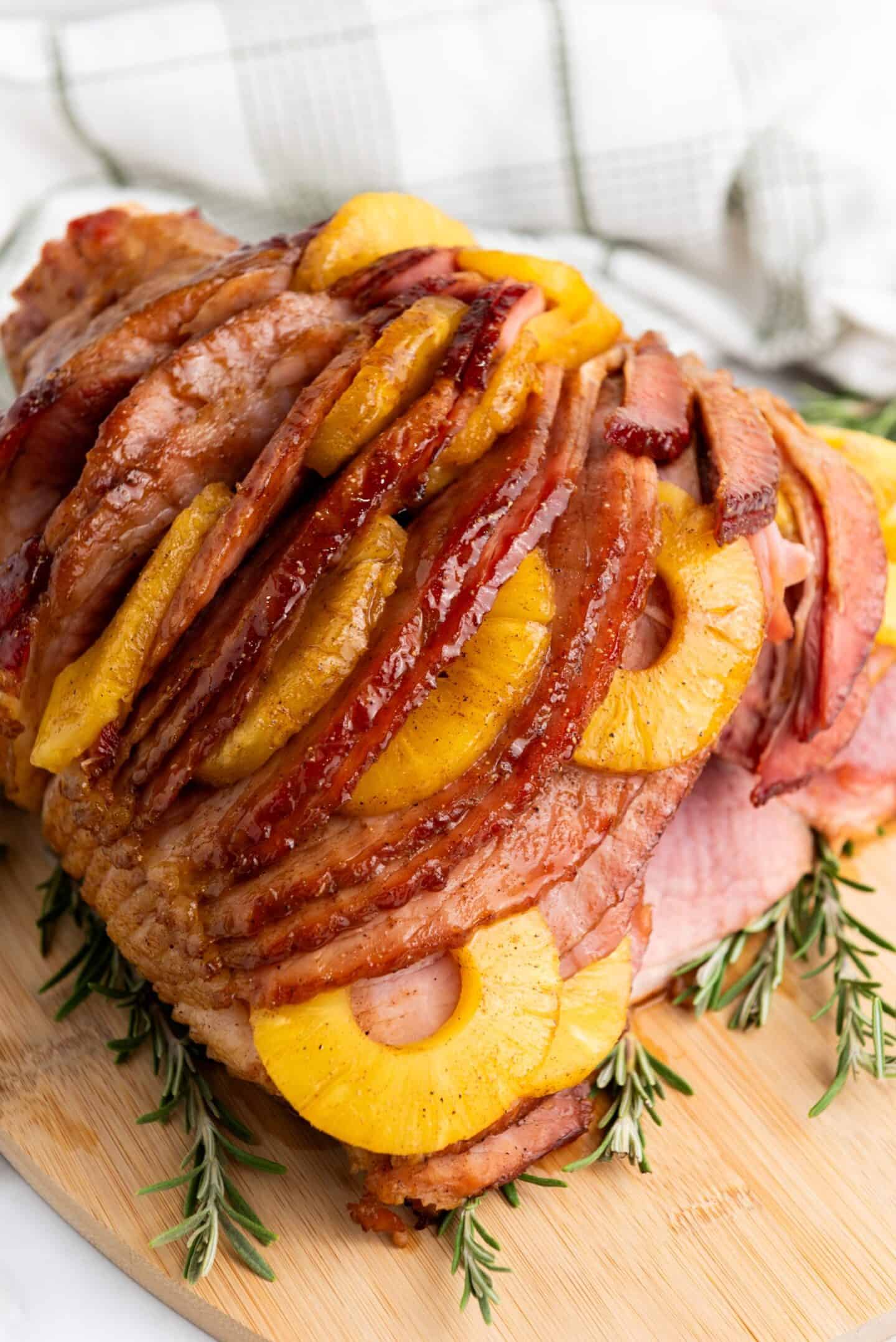 Sliced glazed ham garnished with pineapple rings and served on a wooden board with sprigs of rosemary—perfect for your Easter party food spread.