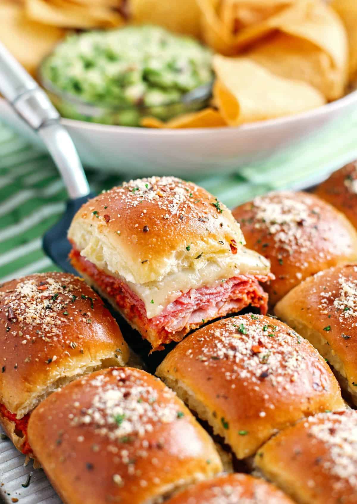 A tray of baked slider sandwiches filled with slices of meat, cheese, and sauce, topped with grated cheese—perfect for an Italian potluck—with chips and guacamole in the background.