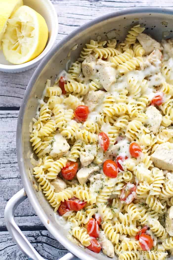 A skillet filled with rotini pasta, pieces of chicken, cherry tomatoes, and melted cheese—perfect for your Spring Dinner Party. A small bowl with lemon wedges sits next to the pan, adding a zesty touch to your dinner party menu.