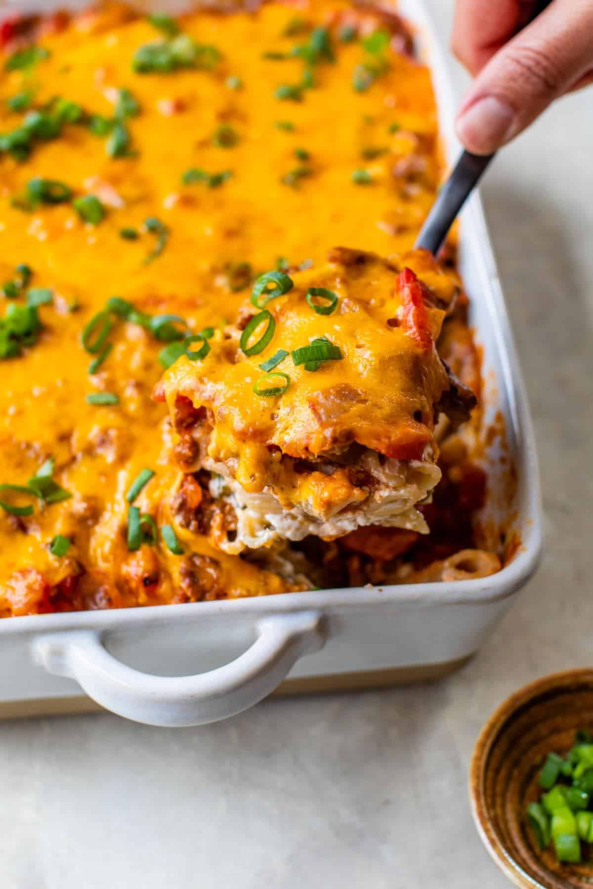 A hand serves a cheesy ground beef dinner casserole topped with chopped green onions from a white baking dish—perfect for easy family dinner ideas.