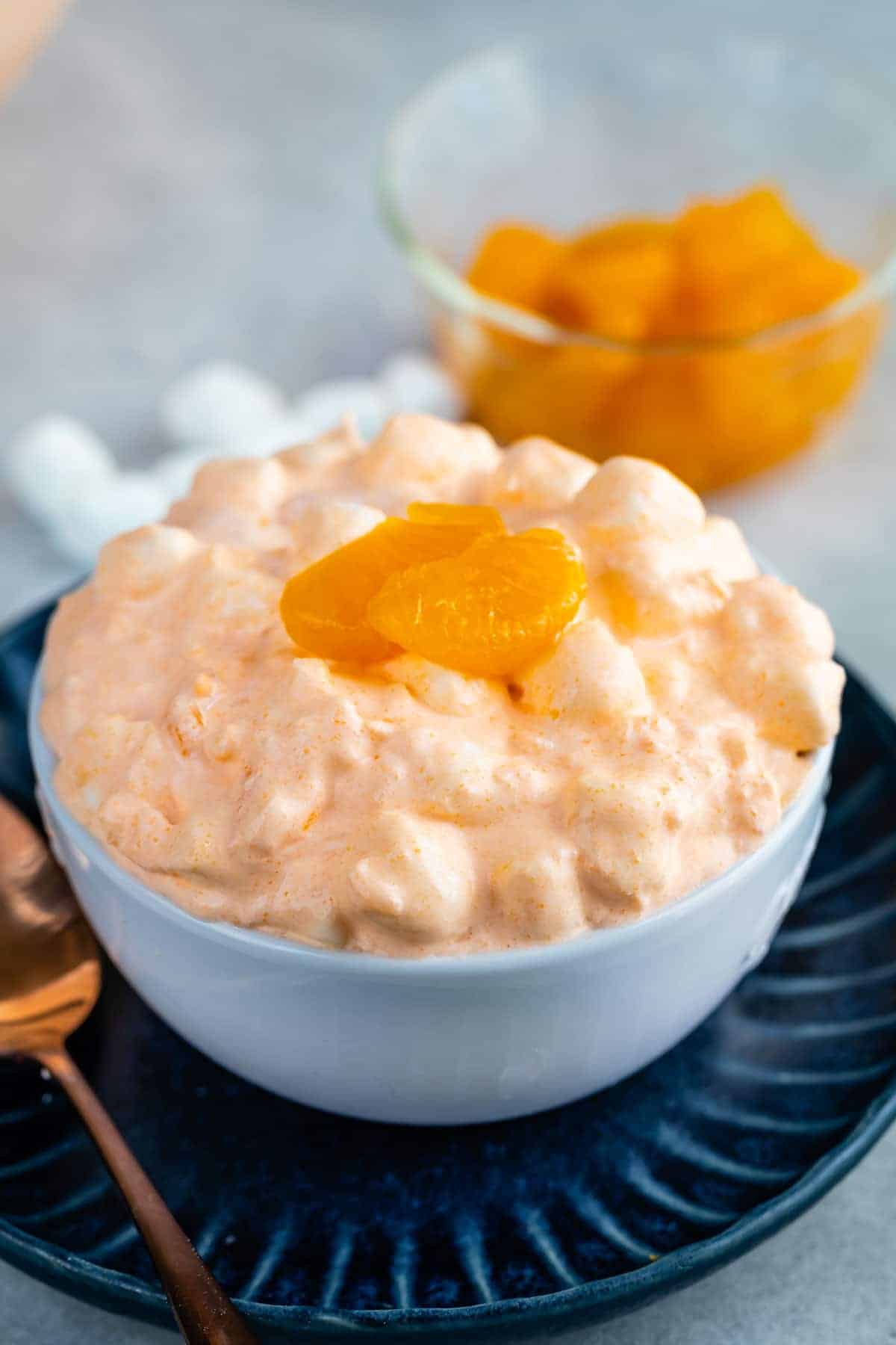 A bowl of creamy orange fluff dessert with mini marshmallows, perfect for Easter side dishes, topped with mandarin orange slices, sits on a blue plate with a spoon and a bowl of mandarin oranges in the background.