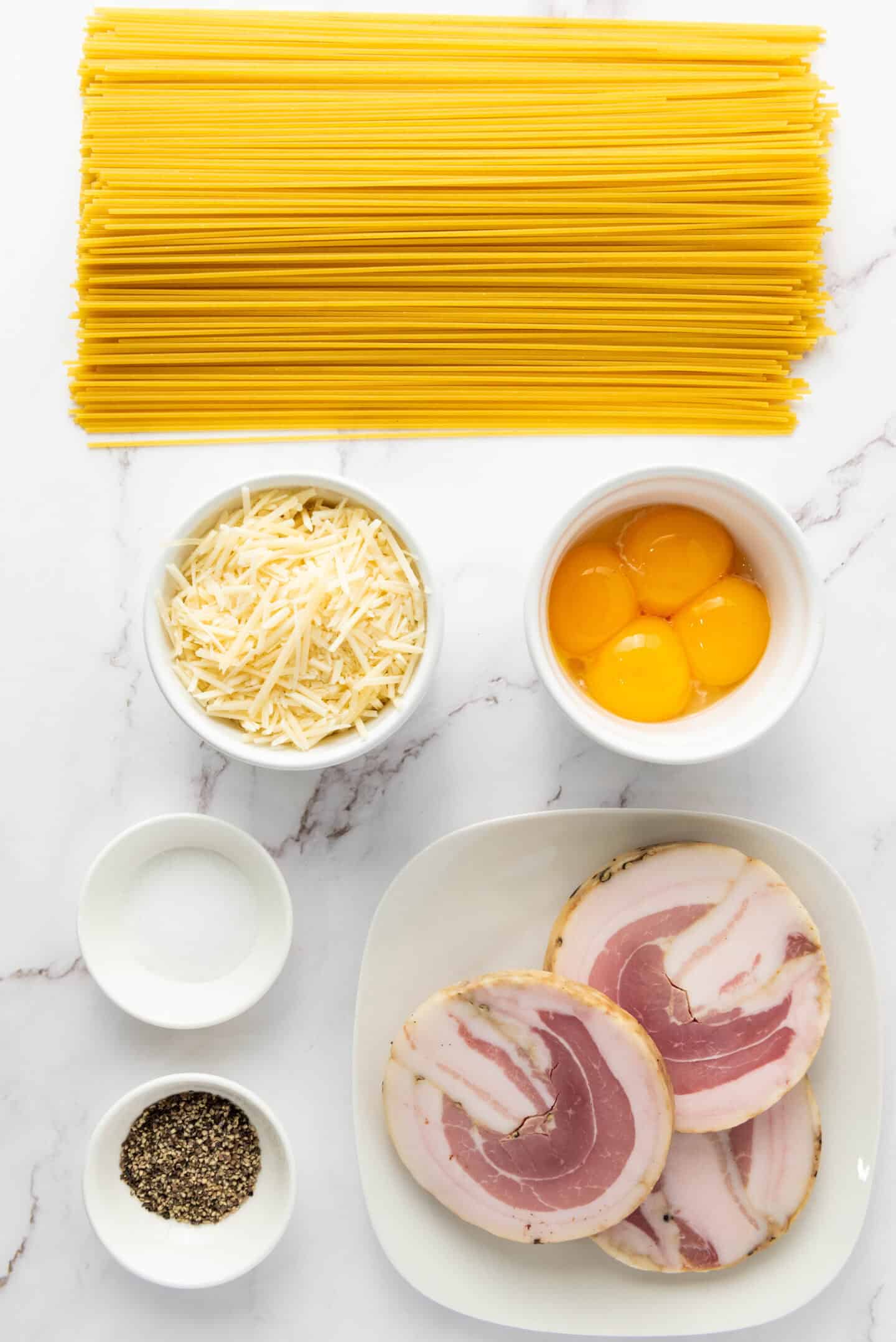 Uncooked spaghetti, grated cheese, egg yolks, pancetta slices, ground black pepper, and salt arranged on a white marble surface—classic ingredients for Authentic Spaghetti Carbonara.