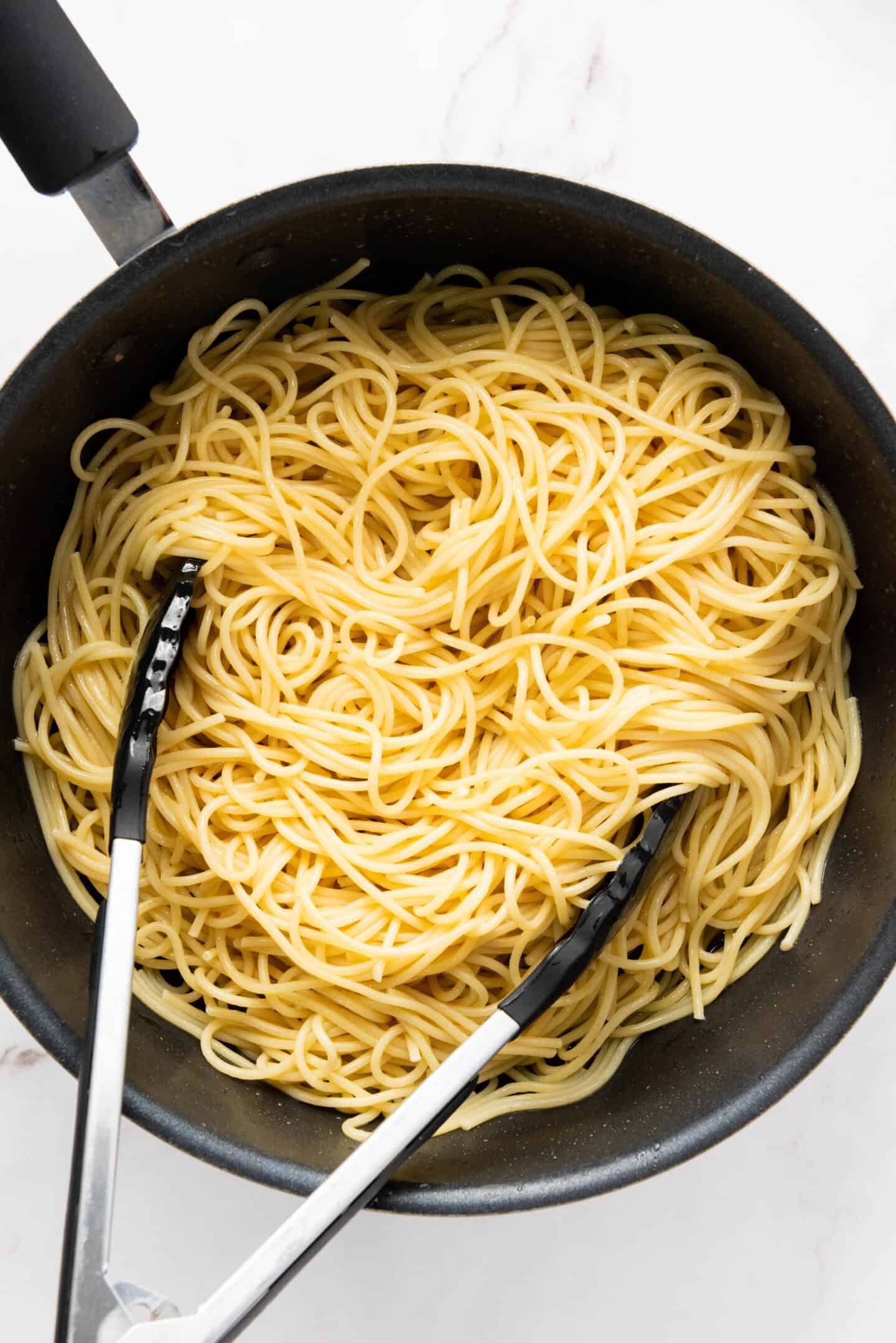 A pot of authentic Spaghetti Carbonara with metal tongs resting on the noodles, placed on a white surface.
