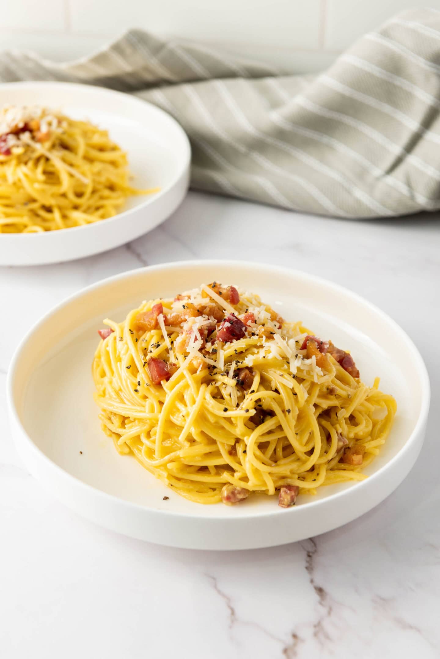 Two white plates with servings of Authentic Carbonara, topped with grated cheese and bacon bits, set on a white marble surface with a striped cloth in the background.
