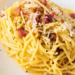 A plate of spaghetti carbonara topped with grated cheese, bacon pieces, and ground black pepper.