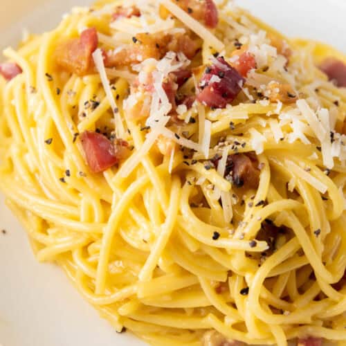 A plate of spaghetti carbonara topped with grated cheese, bacon pieces, and ground black pepper.