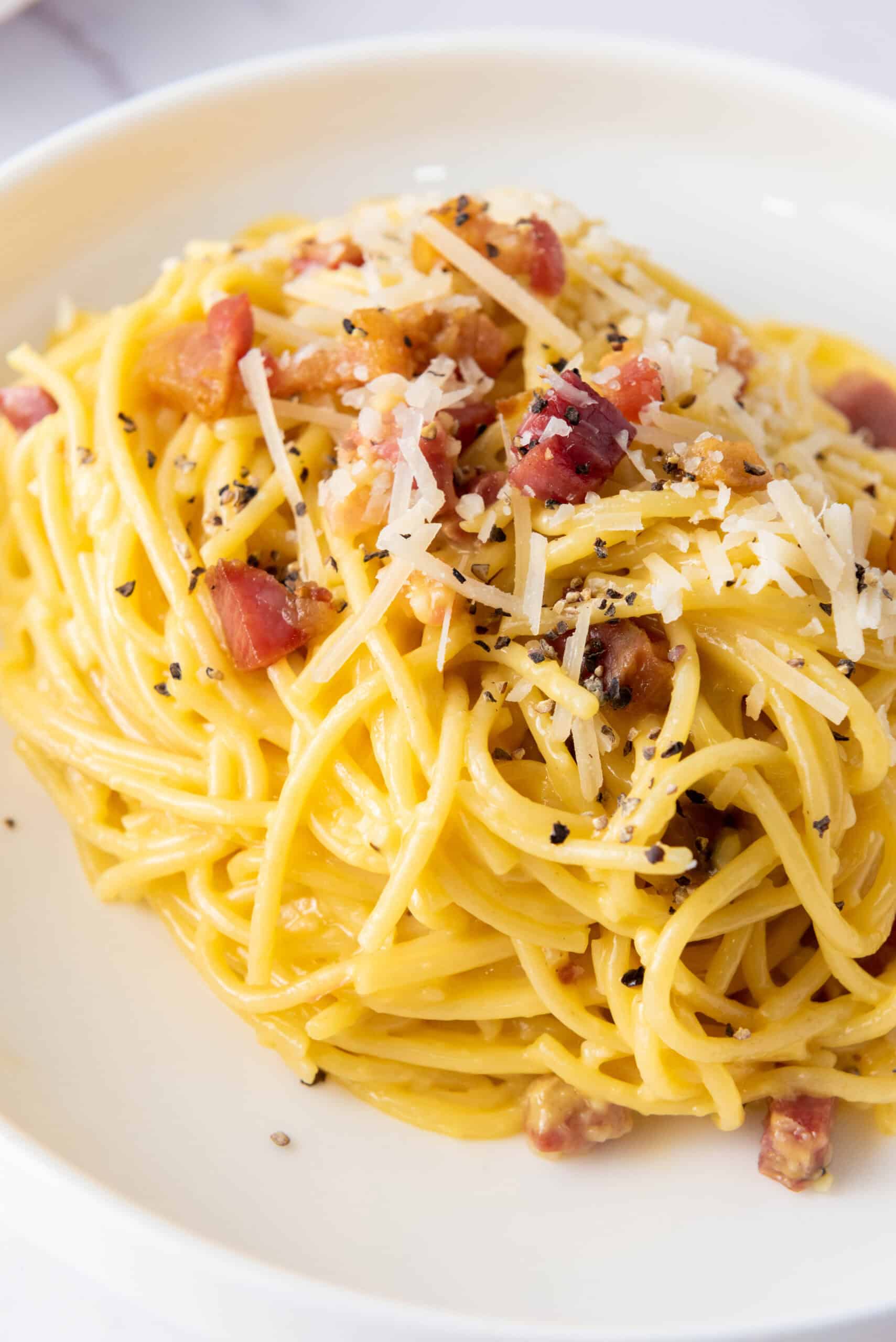 A plate of spaghetti carbonara topped with grated cheese, bacon pieces, and ground black pepper.
