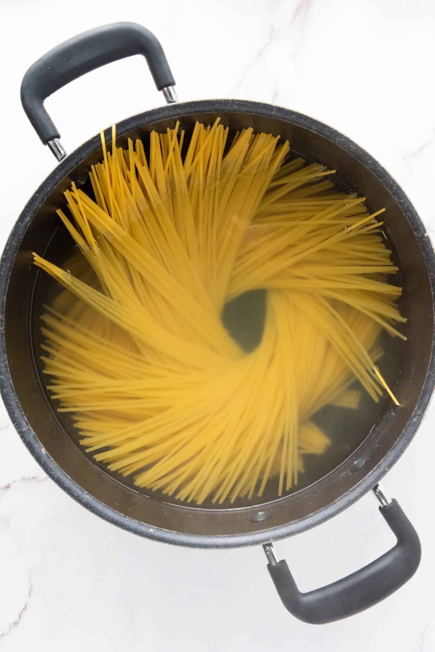 A top-down view of uncooked spaghetti noodles arranged in a spiral pattern in a pot filled with water, ready to be transformed into authentic Spaghetti Carbonara.